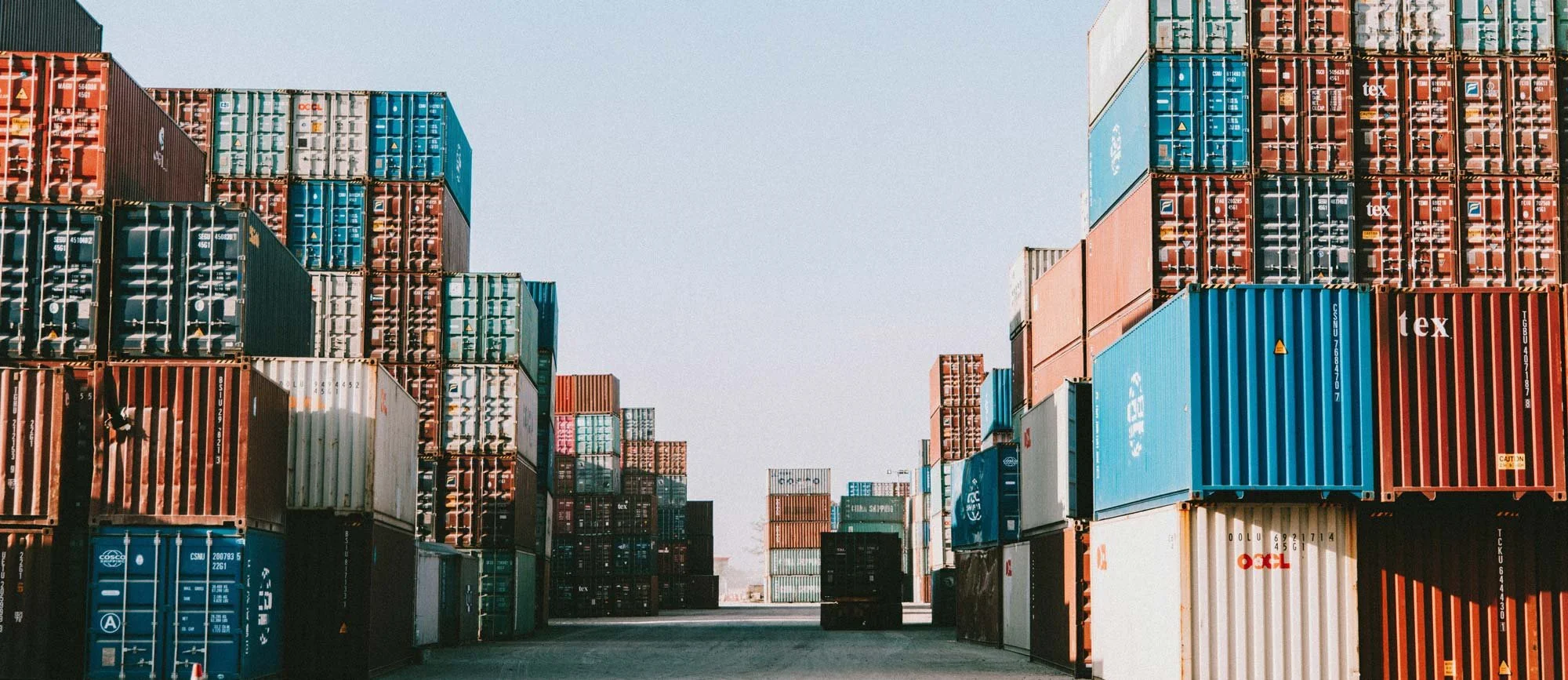 A large outdoor storage area filled with stacked shipping containers of various colors, including red, blue, white, and black, under a clear sky.