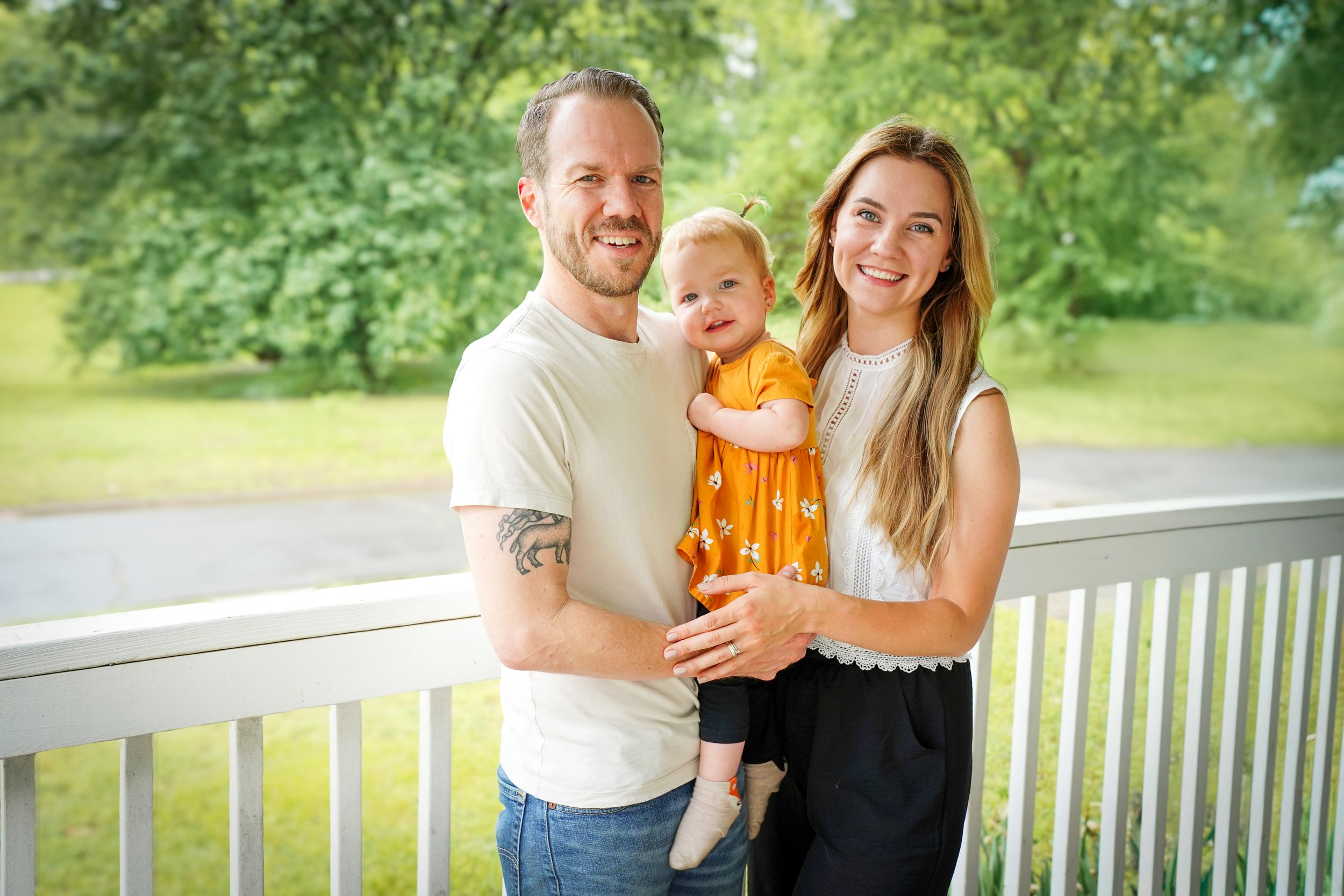 A happy family of three standing outdoors on a porch, with a background of green trees. The father has a beard and tattoos on his arm, the mother has long blonde hair, and they are holding their young daughter who is wearing an orange dress with white flowers.
