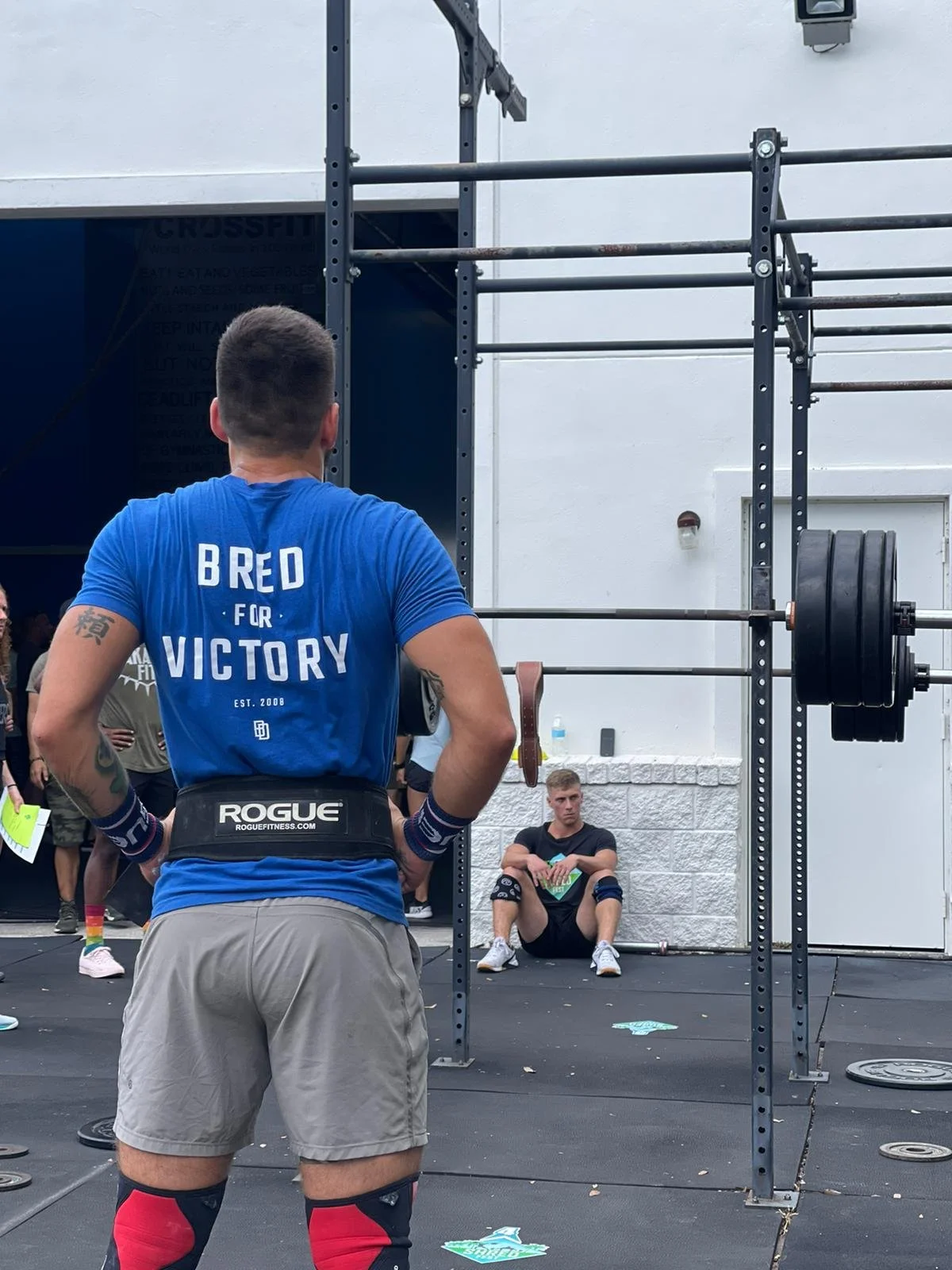A man wearing a blue shirt and gray shorts standing with his back to the camera, observing a young man sitting against a white wall. The young man is in athletic gear, including black shorts, a black shirt, and knee sleeves, sitting on the ground with a water bottle nearby. There is a large metal structure with weights attached, indicating a gym or CrossFit setting.