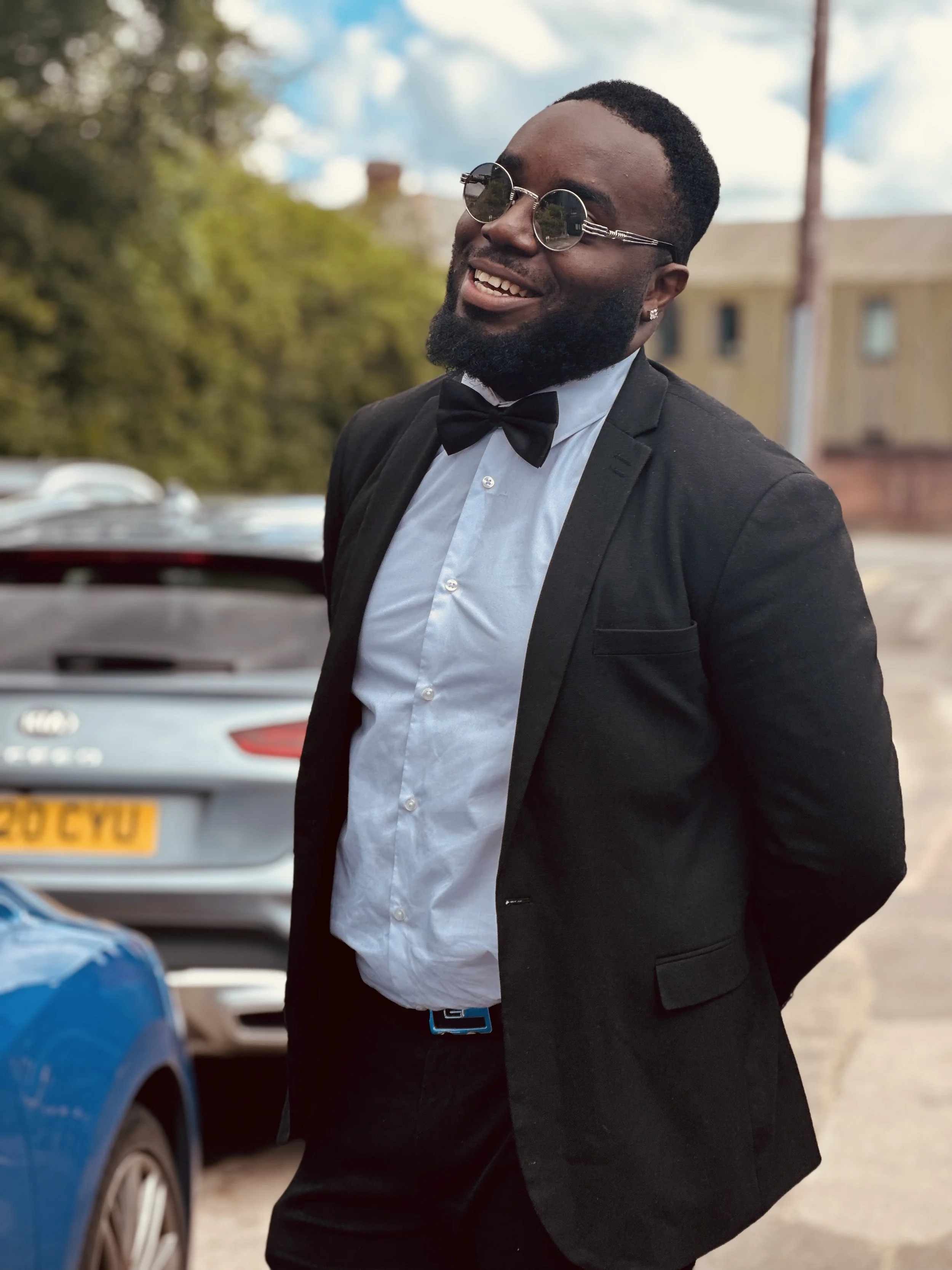 A man in a tuxedo, white shirt, black bow tie, and dark sunglasses is smiling outdoors. He's standing in front of parked cars and a background of trees and buildings.