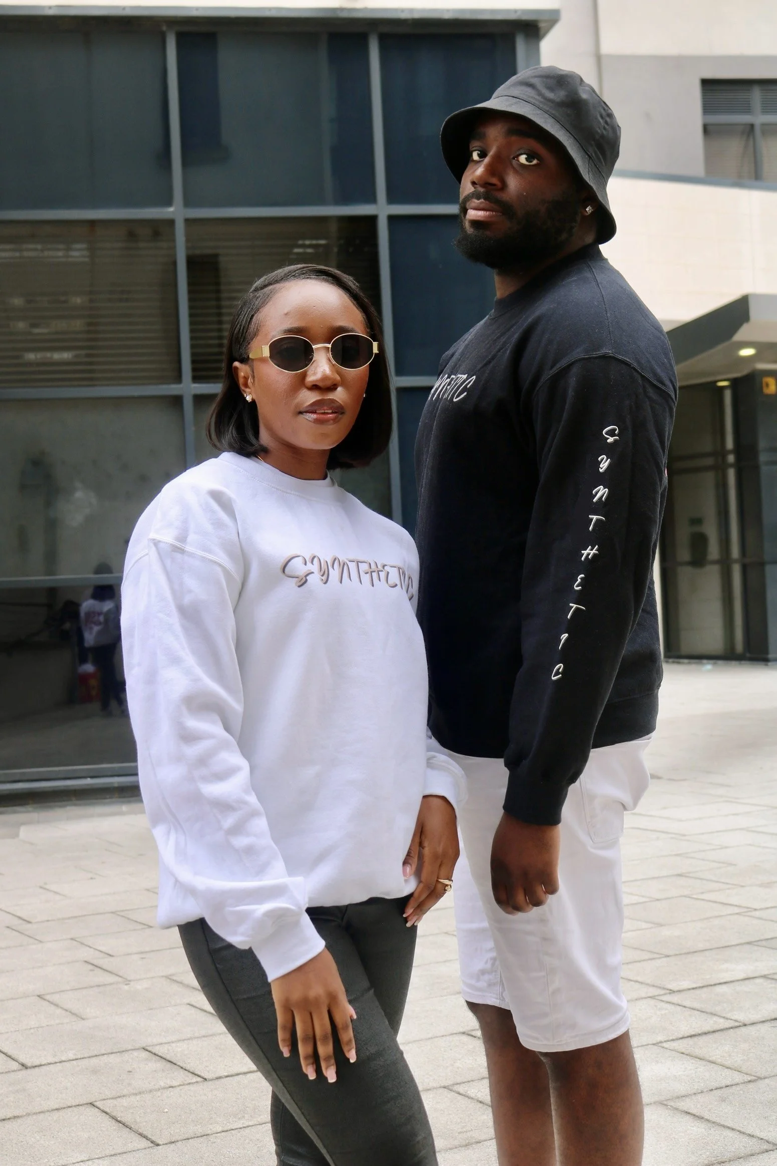 Two people standing outside in an urban setting. The woman on the left is wearing sunglasses, a white sweatshirt with the word 'SYNTHETIC' on it, and dark pants. The man on the right is wearing a black bucket hat, a black sweatshirt with the word 'SYNTHETIC' on the sleeve, and light-colored shorts. They are posed closely with neutral expressions.