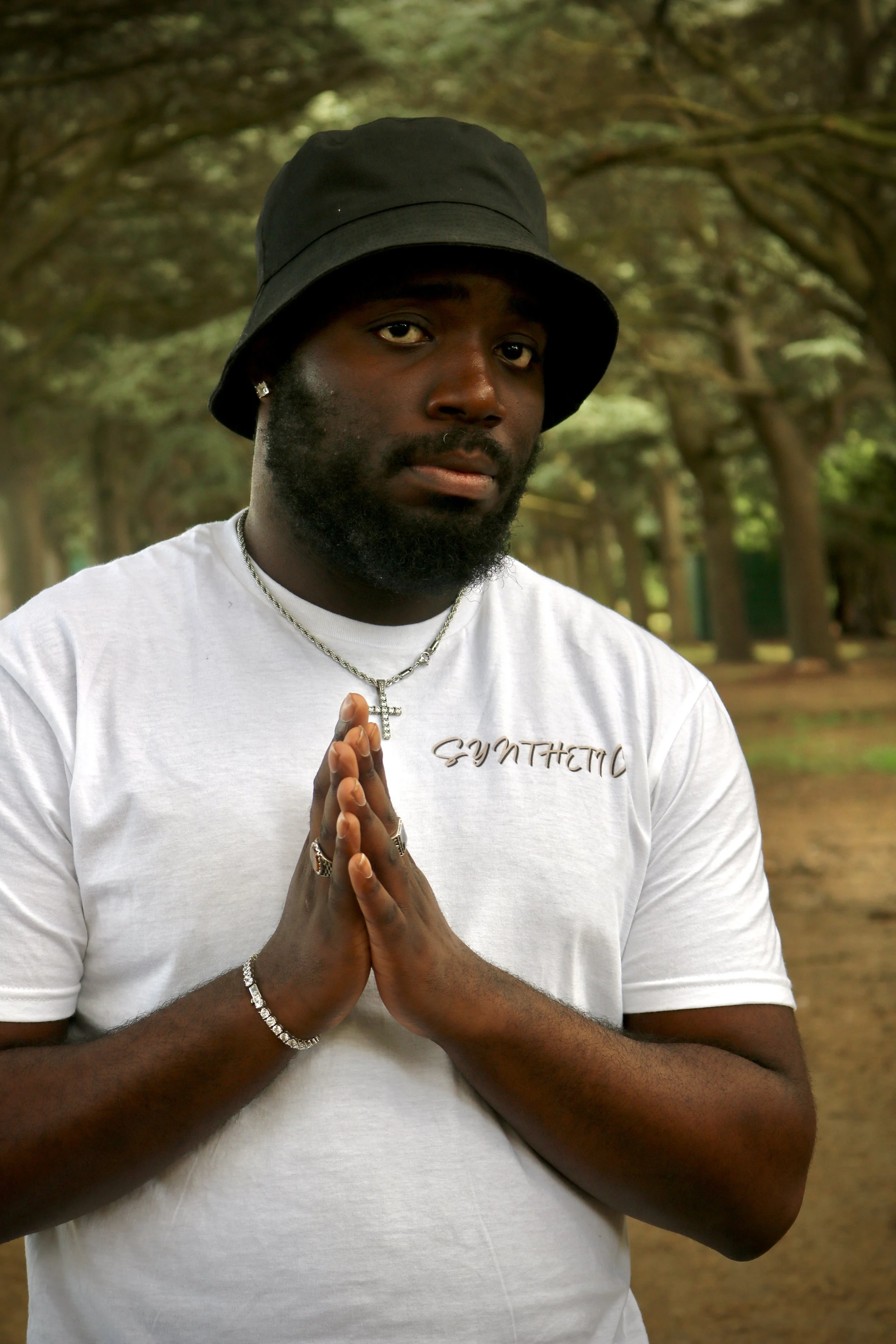 A man with dark skin and a beard wears a black bucket hat and a white t-shirt with the word 'SYNTHETIC' written on it. He has jewelry including a cross necklace, rings, and a bracelet. He is standing outdoors on a wooded path with trees in the background, holding his hands together in a prayer-like gesture.