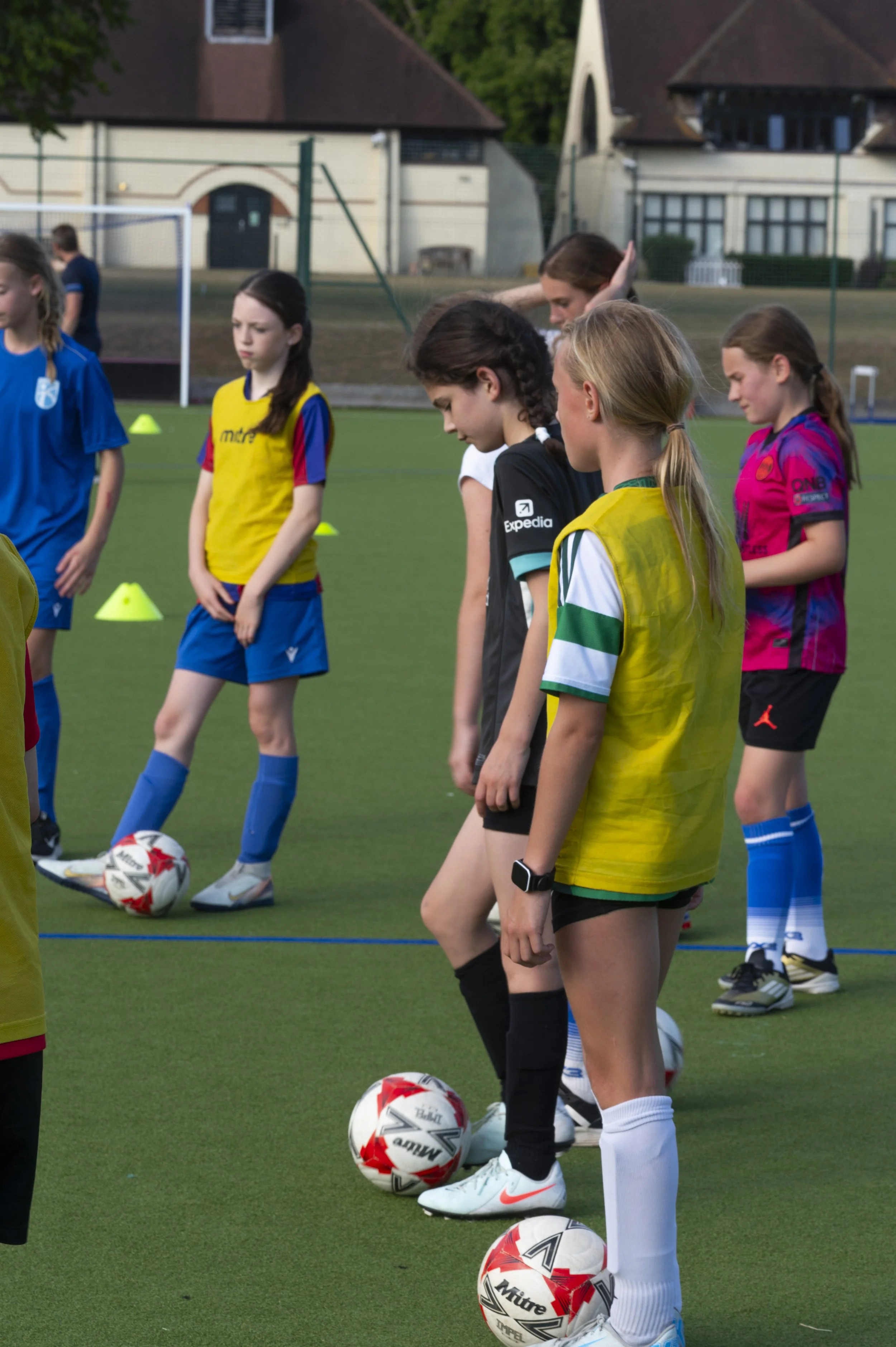 Young female soccer players standing on a field with soccer balls, preparing for a practice or game, surrounded by training cones.