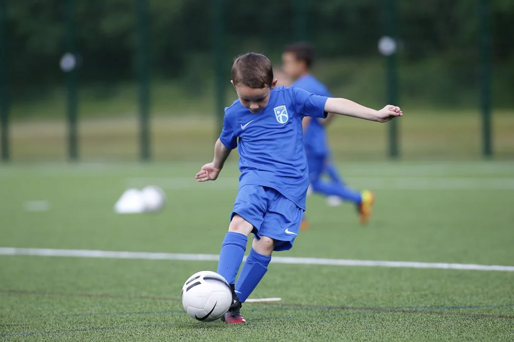 Young boy in blue soccer uniform kicking a soccer ball on a field, with another child in the background.