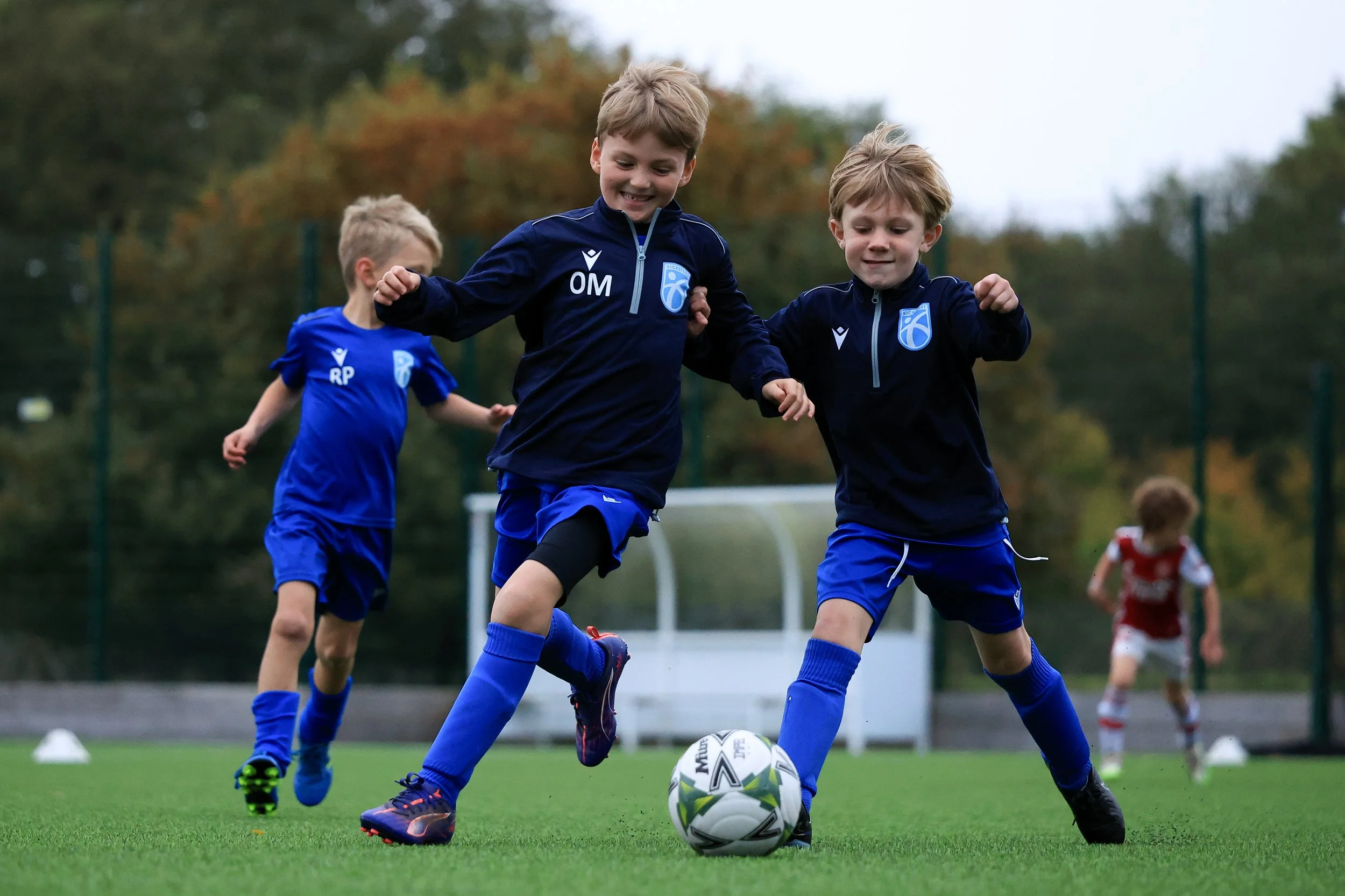 Young boys playing soccer on a field, with two in the foreground fighting for the ball, and two others in the background, fall and autumn trees visible.