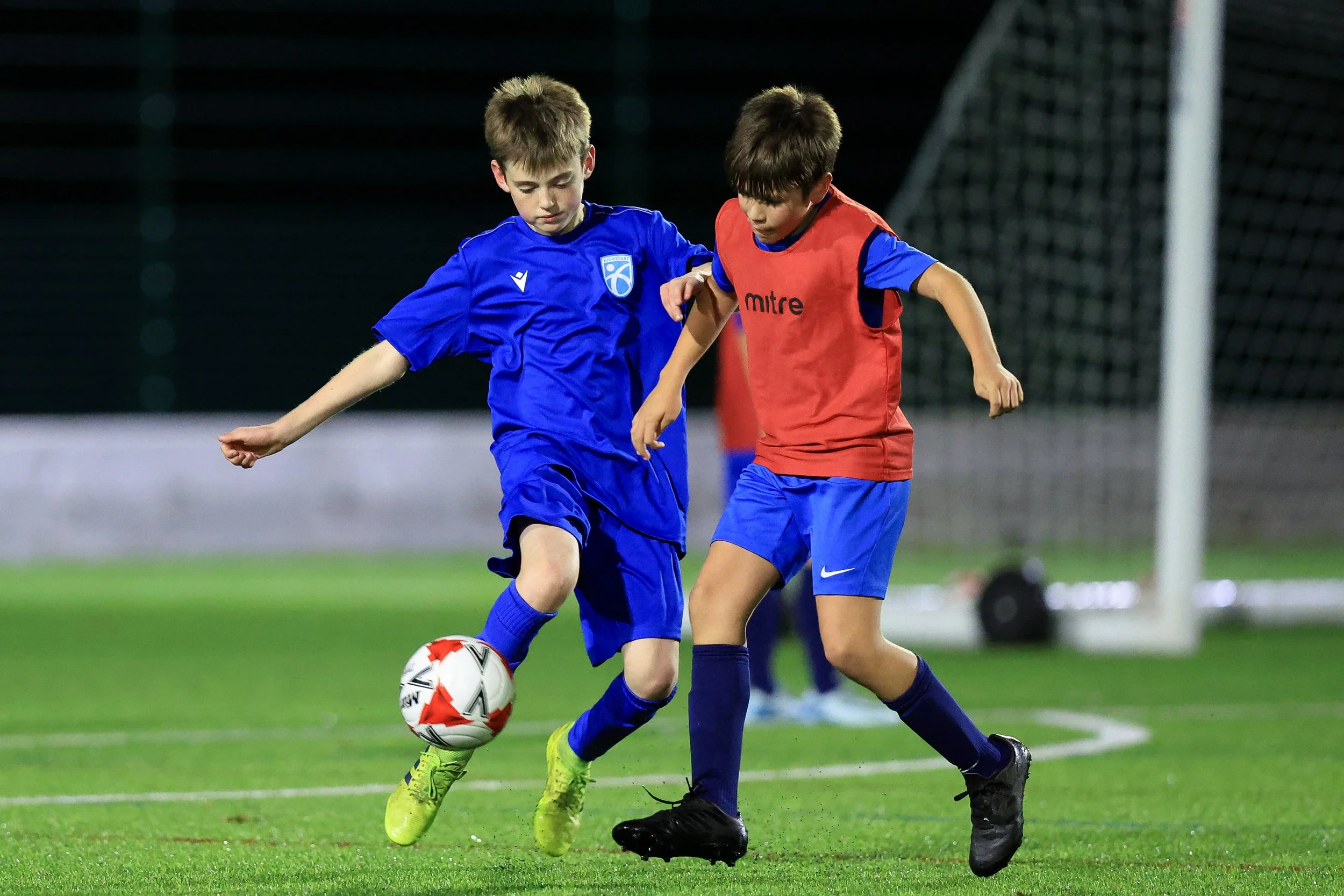 Two young boys playing soccer on a field at night, wearing blue and red sports uniforms, focusing on the soccer ball.