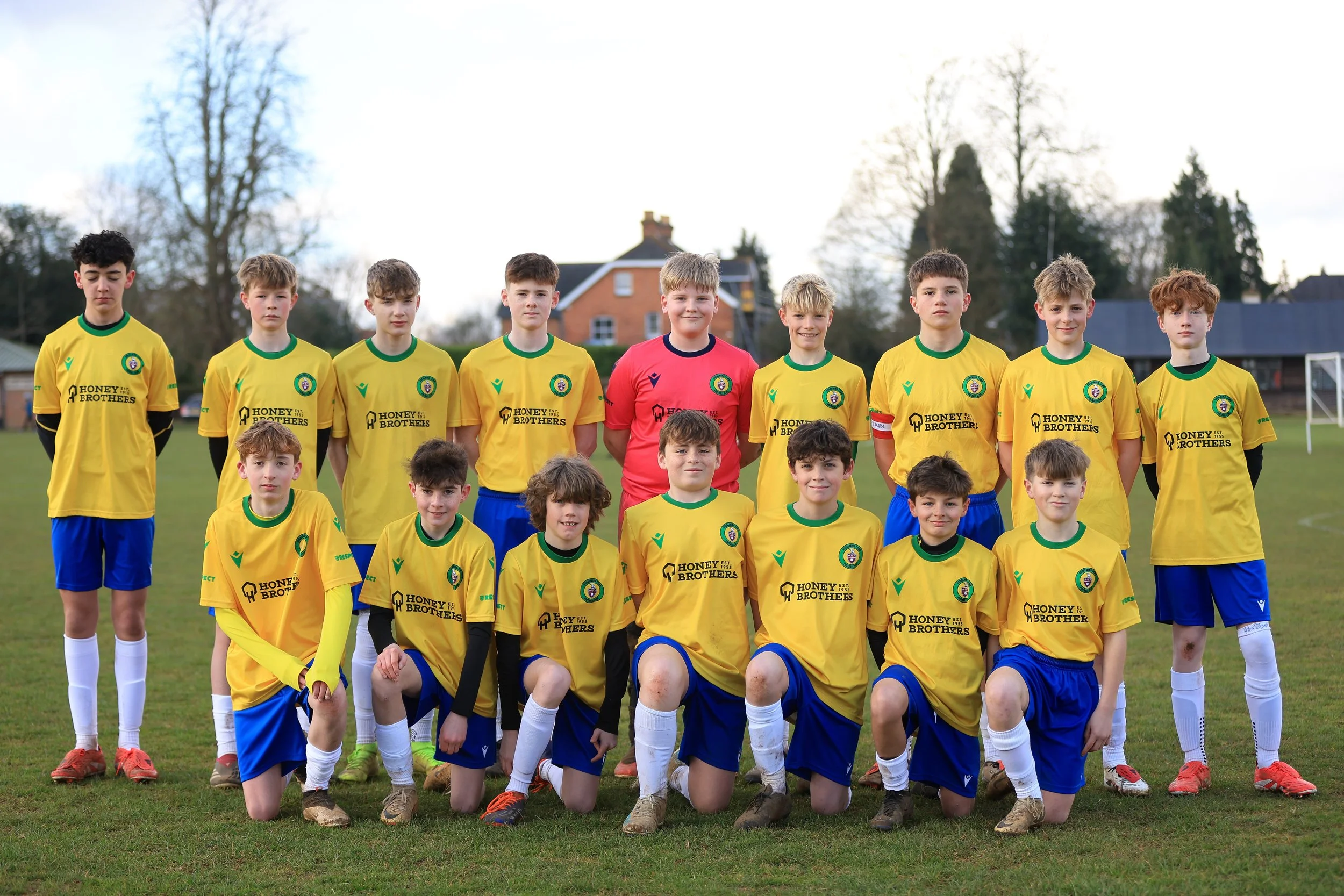 A youth soccer team of 14 boys posing on a grass field, wearing yellow jerseys with blue shorts and white socks, with a blonde goalkeeper in a red jersey in the center of the back row, during daytime.