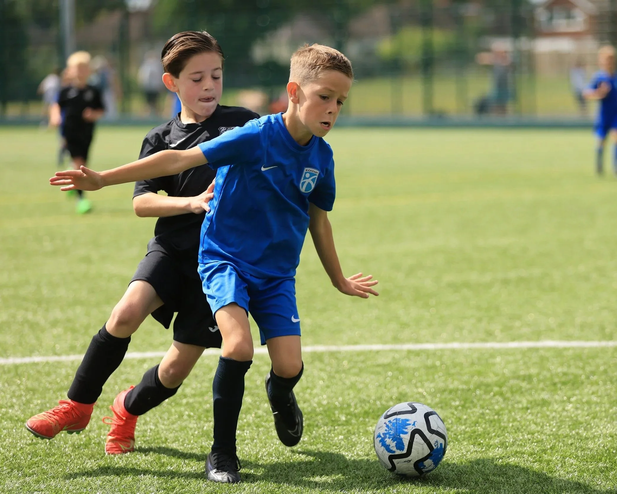 Two boys playing soccer on a grassy field, one in a blue jersey and the other in a black jersey, chasing a soccer ball.