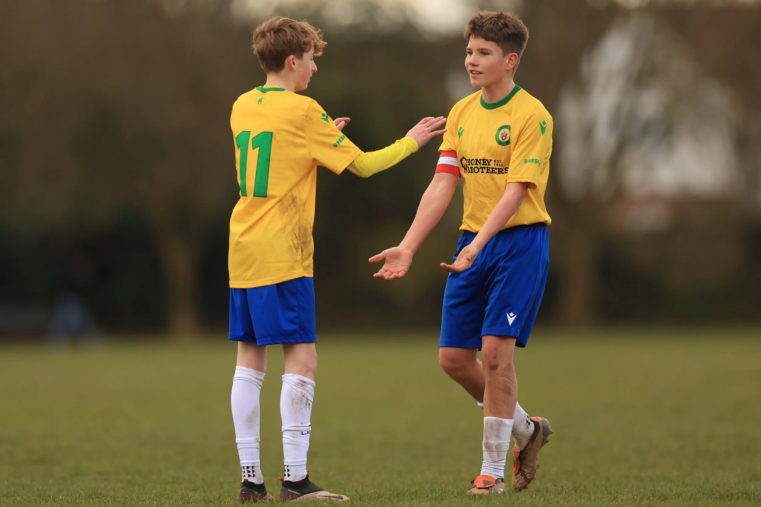 Two young boys with soccer uniforms on a field, one with jersey number 11 and the other' with a captain's armband, appear to be engaged in a conversation or celebration.