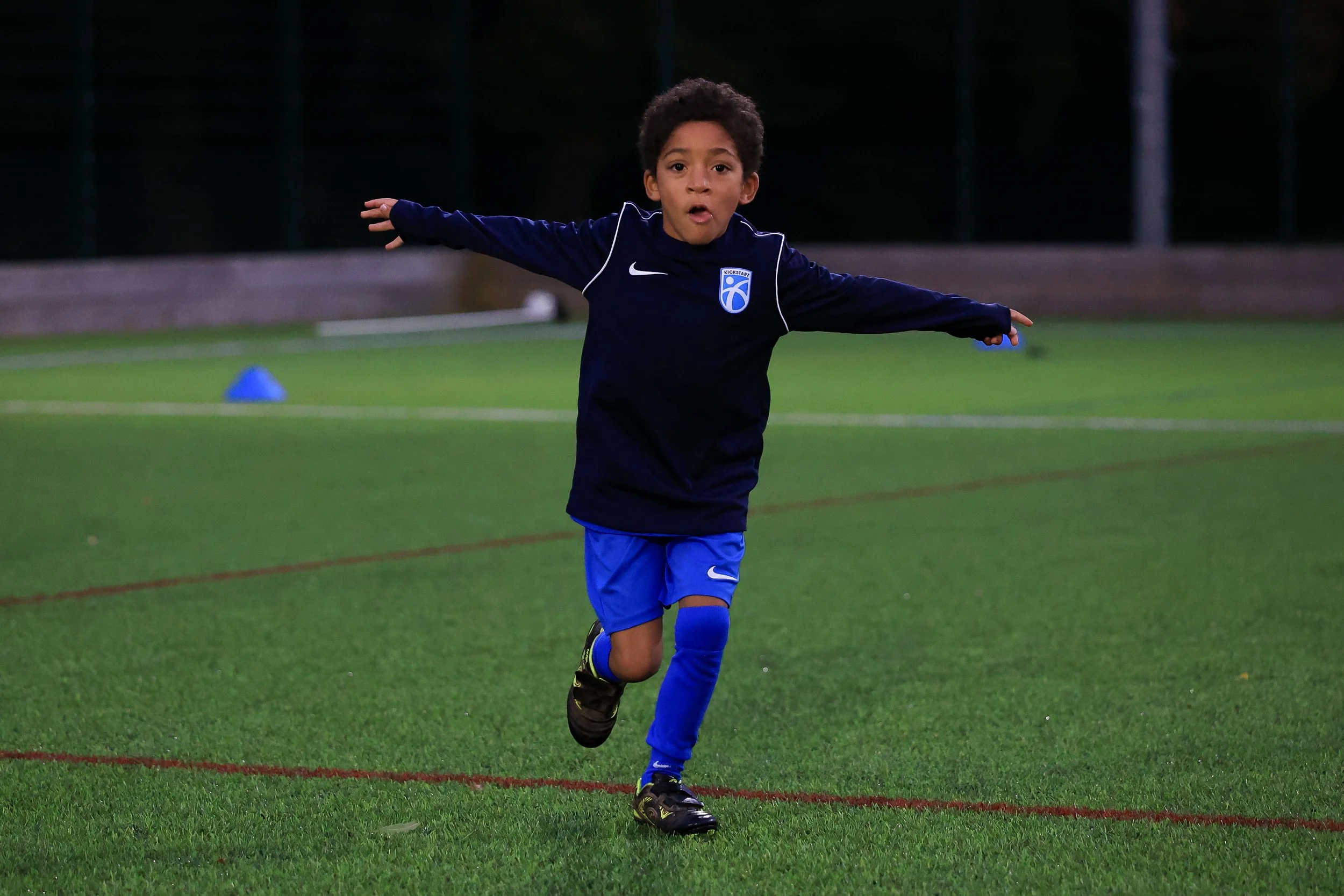 A young boy in a black and blue soccer uniform running on a field with arms outstretched, playing soccer during evening.