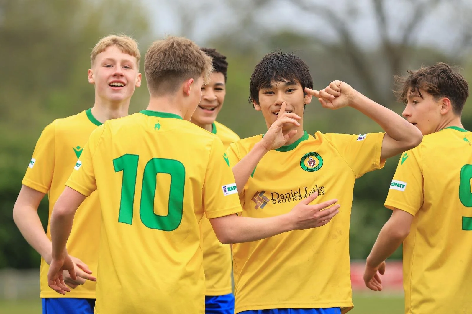 Young soccer players in yellow and green uniforms celebrate on the field, one making a gesture near his ear while the others smile and interact, with trees in the background.