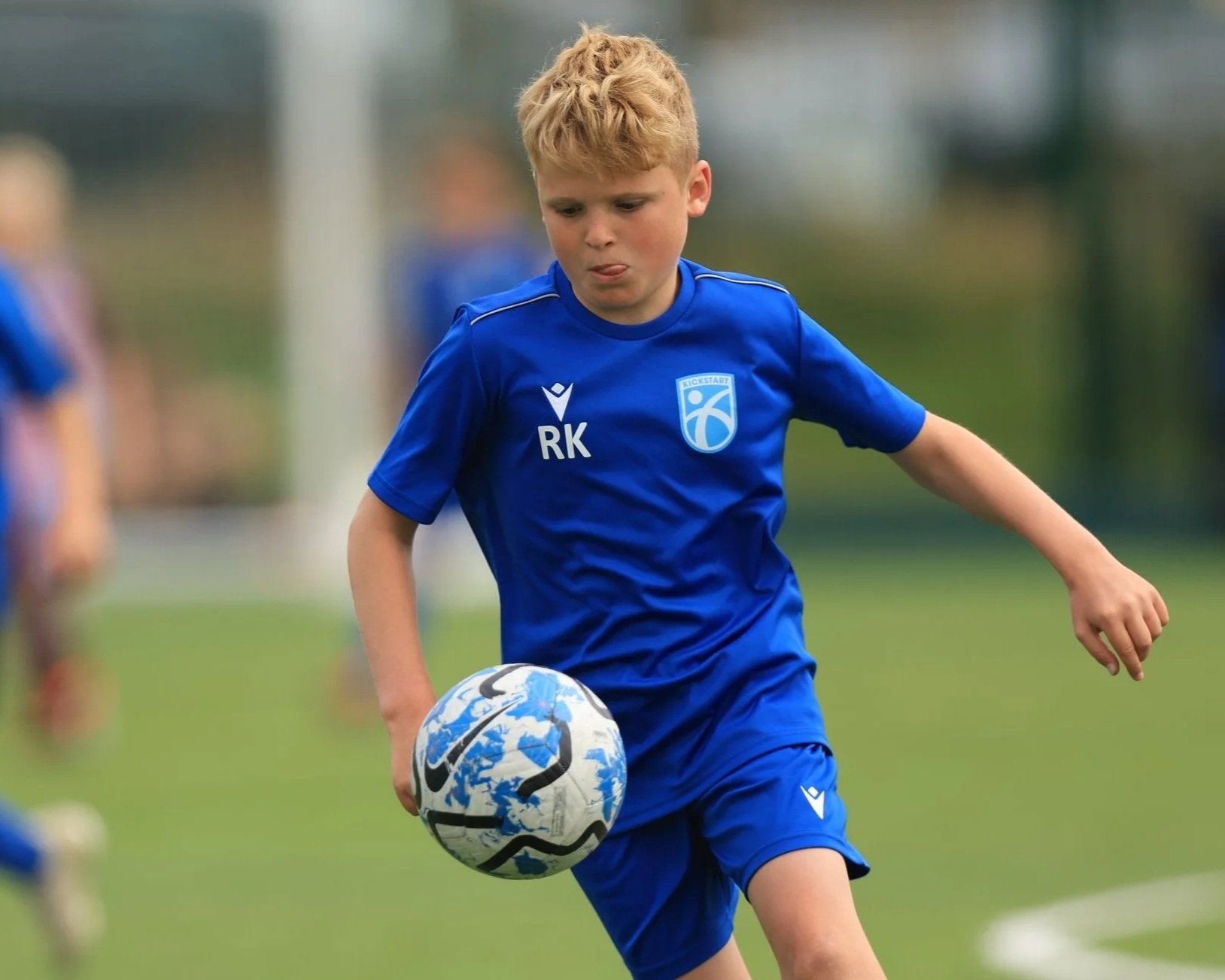 A young boy with curly blond hair wearing a blue sports uniform with the logo 'Kickstart' and initials 'RK', playing soccer outdoors on a grassy field, controlling a soccer ball with his right foot.