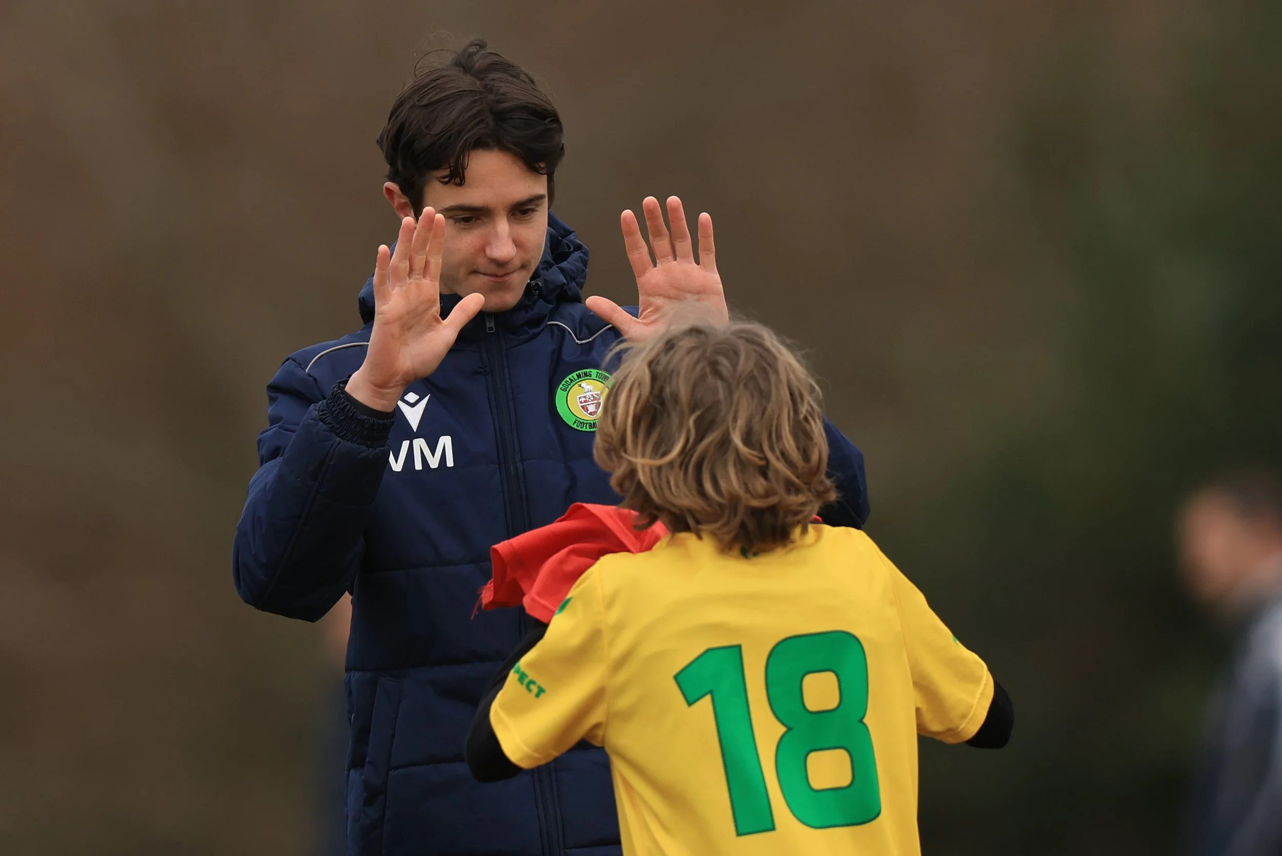 A young man in a navy blue jacket with a green, red, and white badge is giving a high five to a young boy with blond curly hair wearing a yellow sports jersey with green numbers and letters. The background is blurred with muted colors.