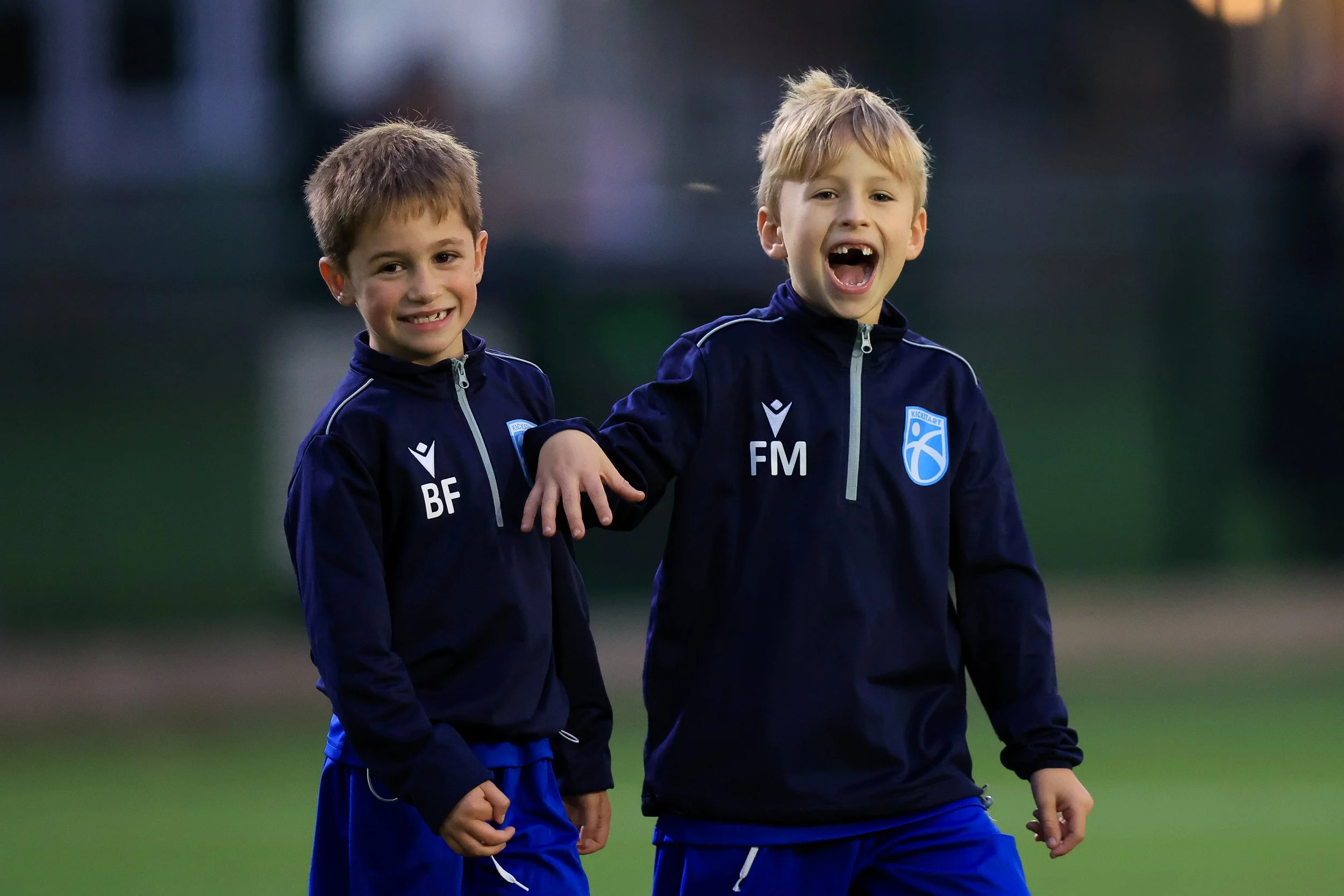 Two young boys in navy sports jackets with blue shorts, smiling and laughing on a sports field during dusk or early evening.