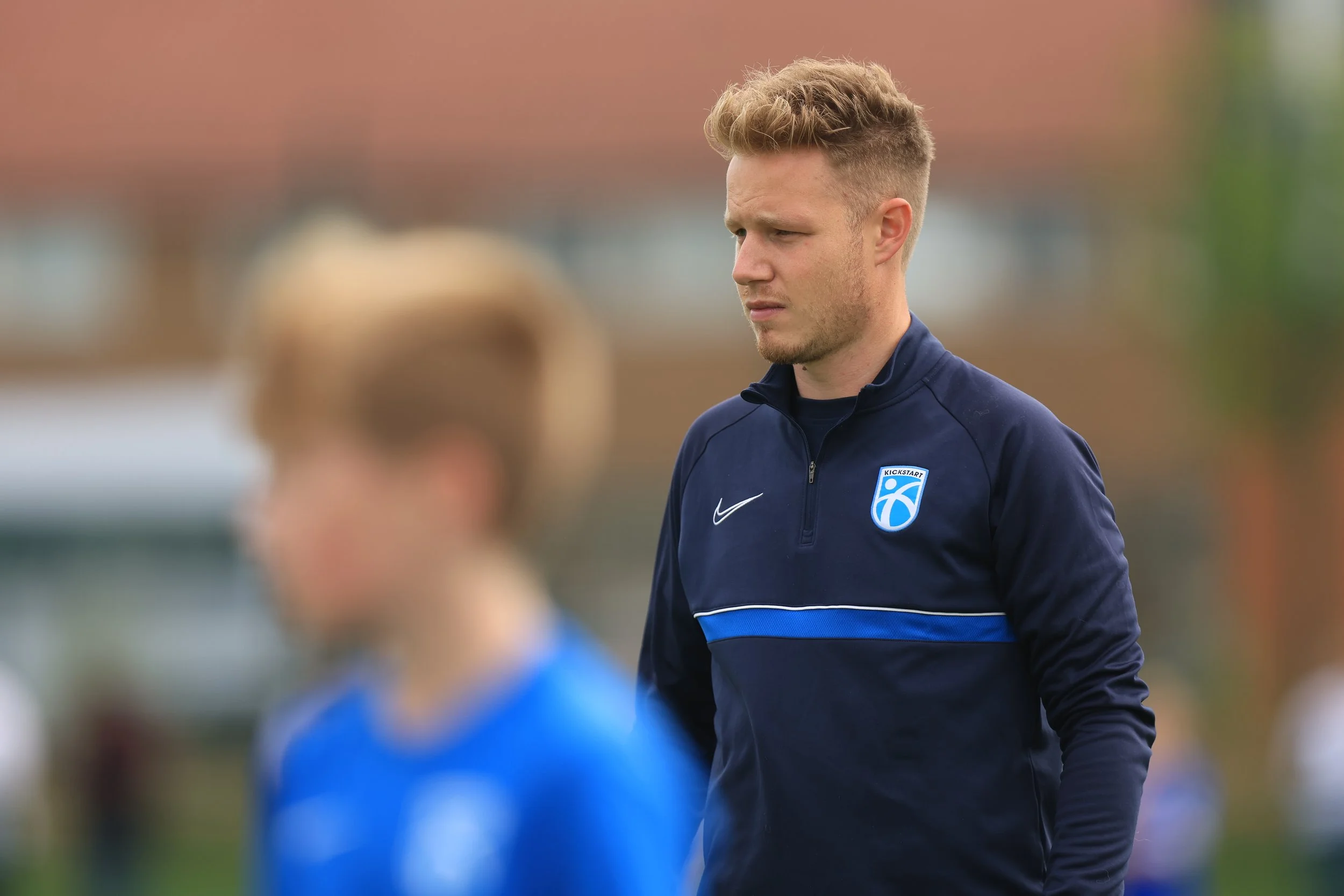 A young man with short, curly blond hair and a light beard wearing a navy blue Nike sports jacket with a logo on it, standing outdoors on a sports field, looking down with a serious expression.