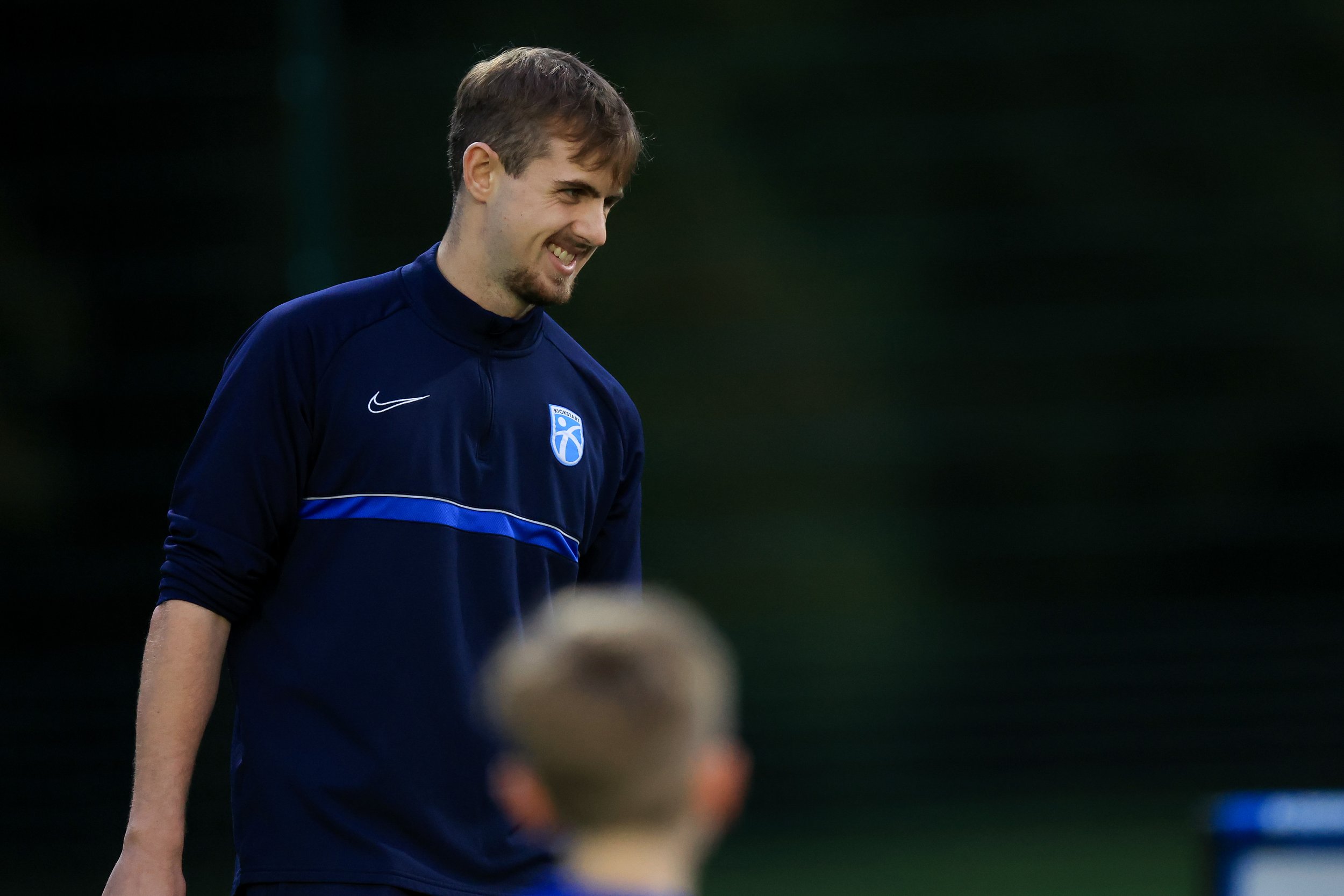 A young man in a navy blue sports jacket with a blue and white emblem on the chest, smiling and looking down during a sports practice or game, with another person blurred in the foreground.