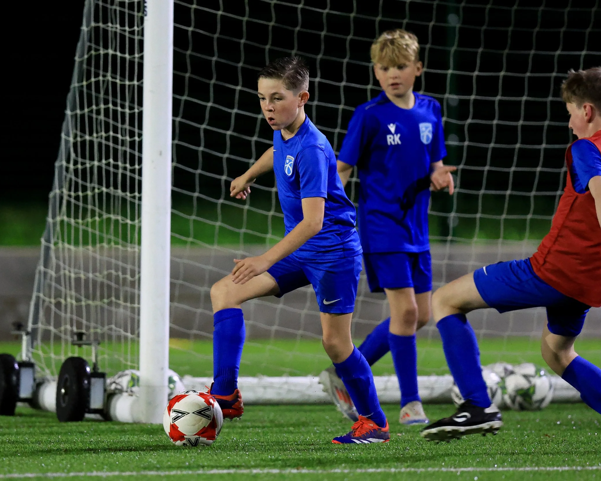 Young boys playing soccer on a field at night, with one player preparing to kick the ball near the goal, and others watching, all wearing blue jerseys.