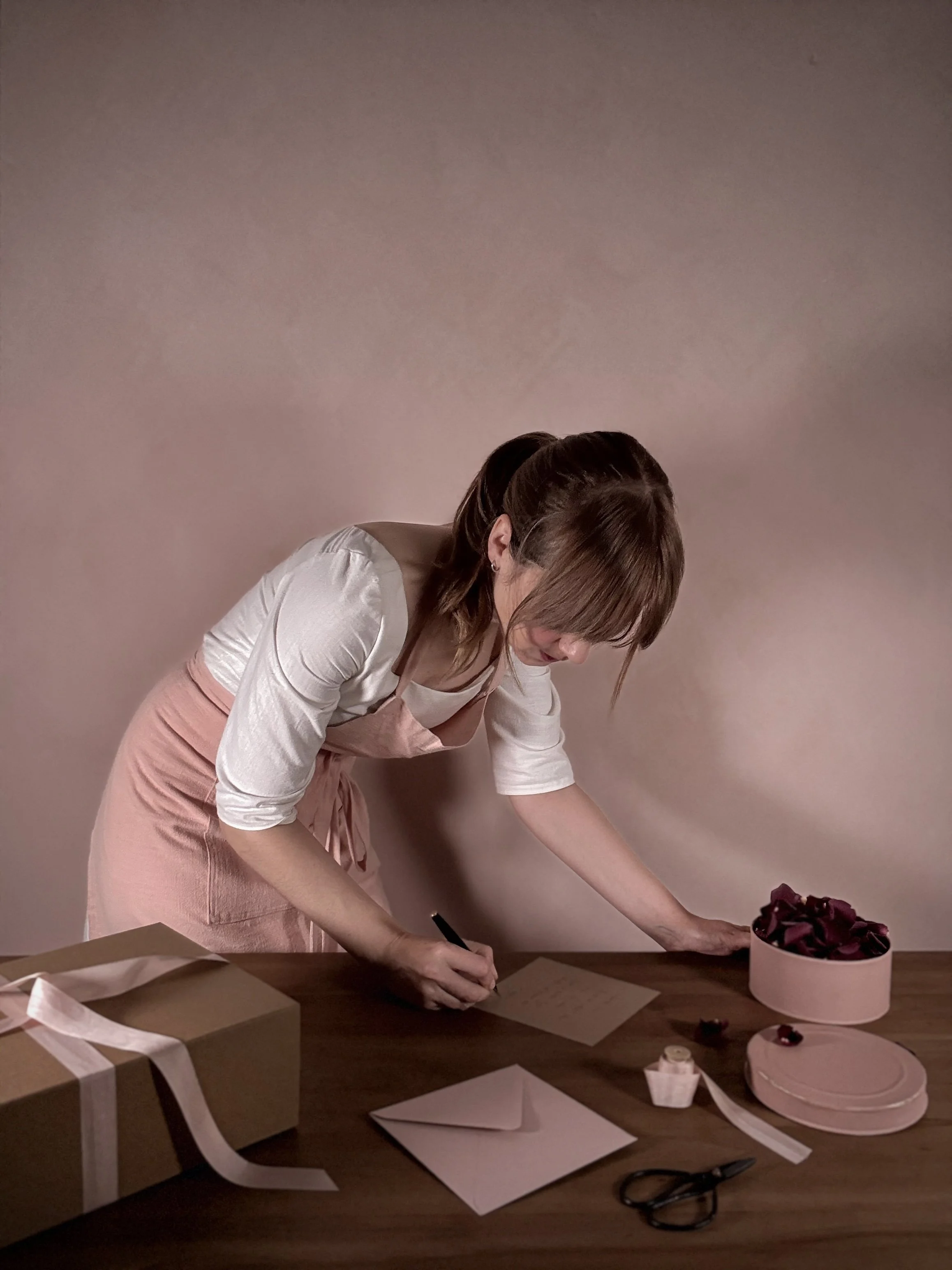 Mujer decorando una caja de regalo con flores rojas junto a otros objetos de envoltorio sobre una mesa.