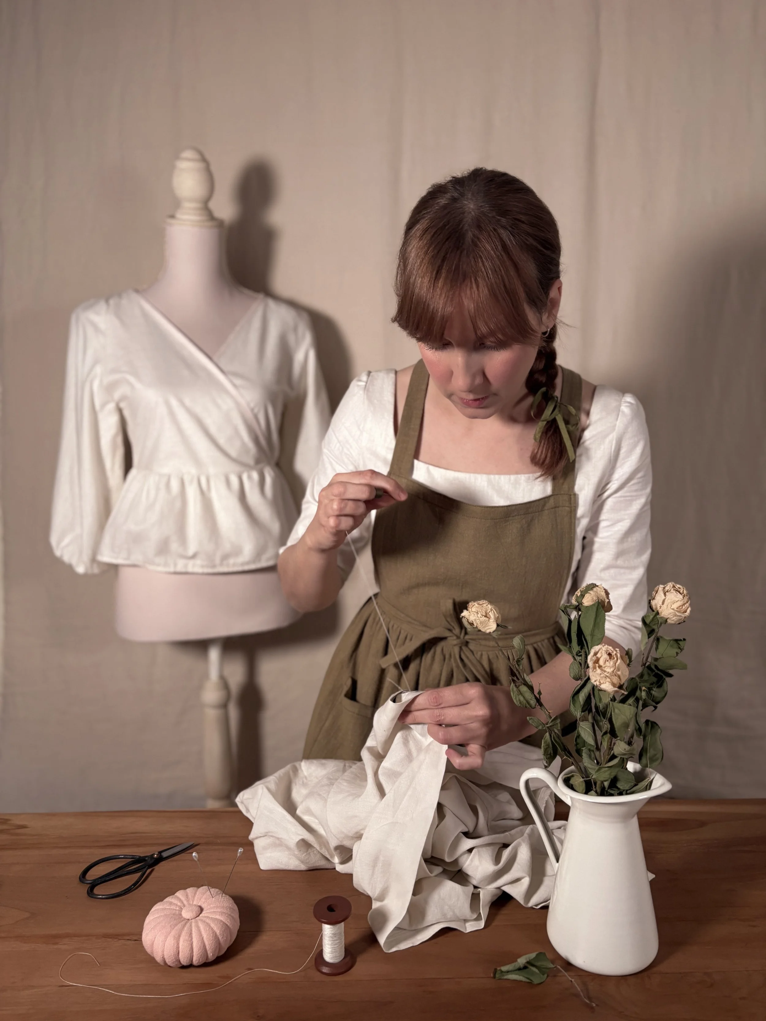 Mujer arreglando un ramo de rosas secas en una mesa de madera, con una tijera, un carrete de hilo y una almohadilla de coser en la mesa, y un maniquí con blusa blanca y falda en el fondo.