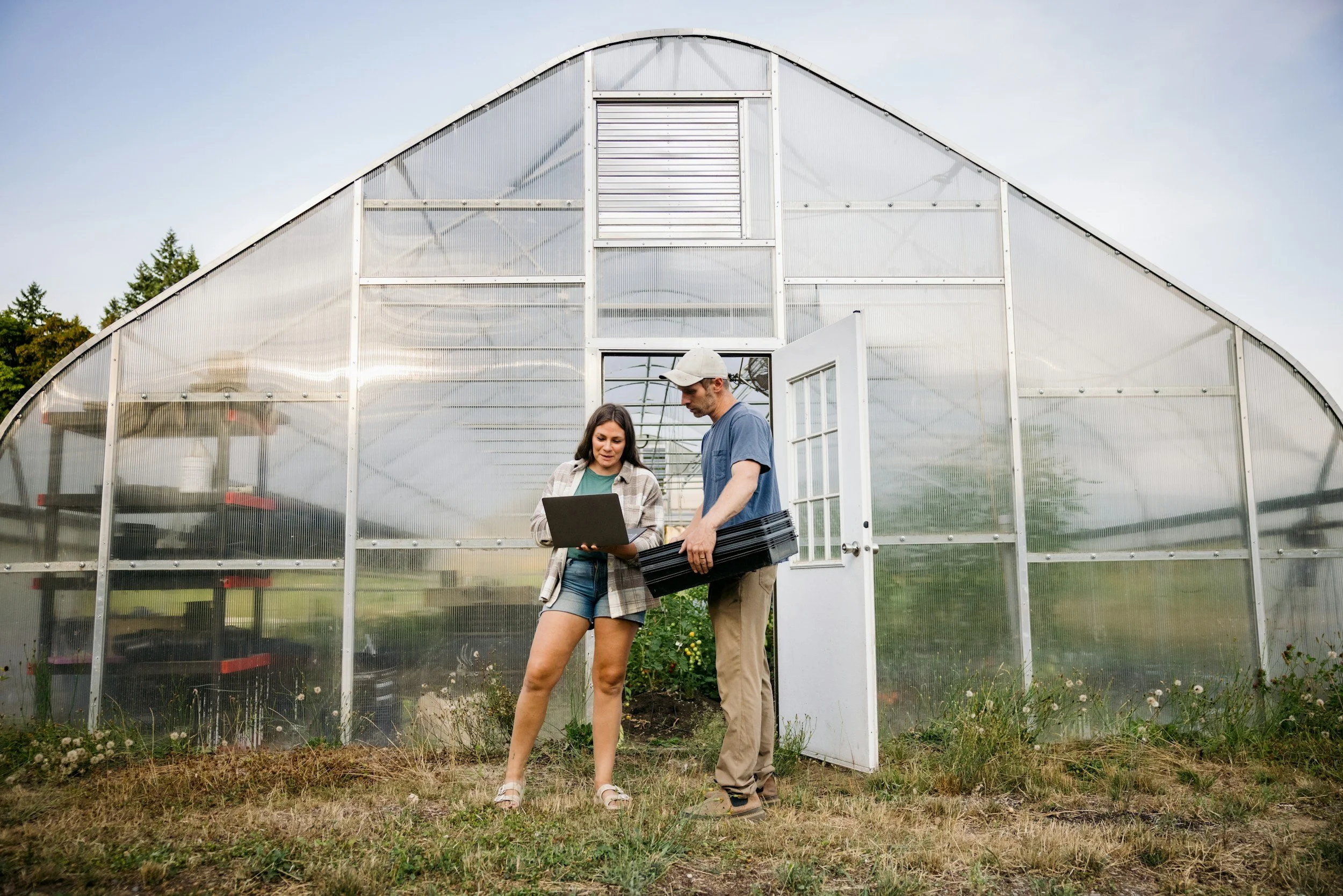 Two people, a man and a woman, standing outside a greenhouse, discussing something on a laptop, with gardening trays in the woman's hands.