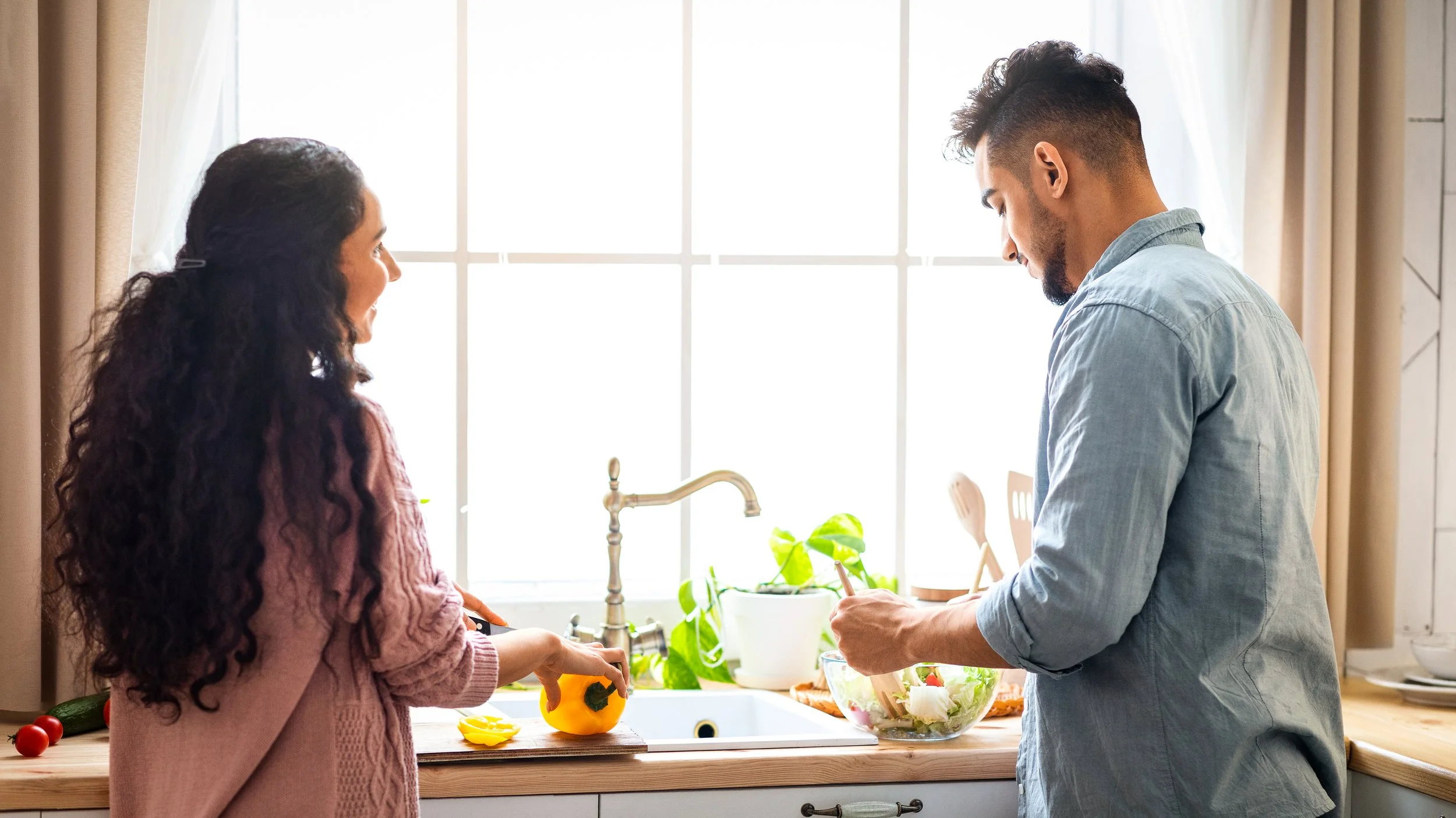 A young man and woman are talking in a bright kitchen, preparing food together. The woman stands by the sink holding a yellow bell pepper, while the man is chopping vegetables in a salad bowl.