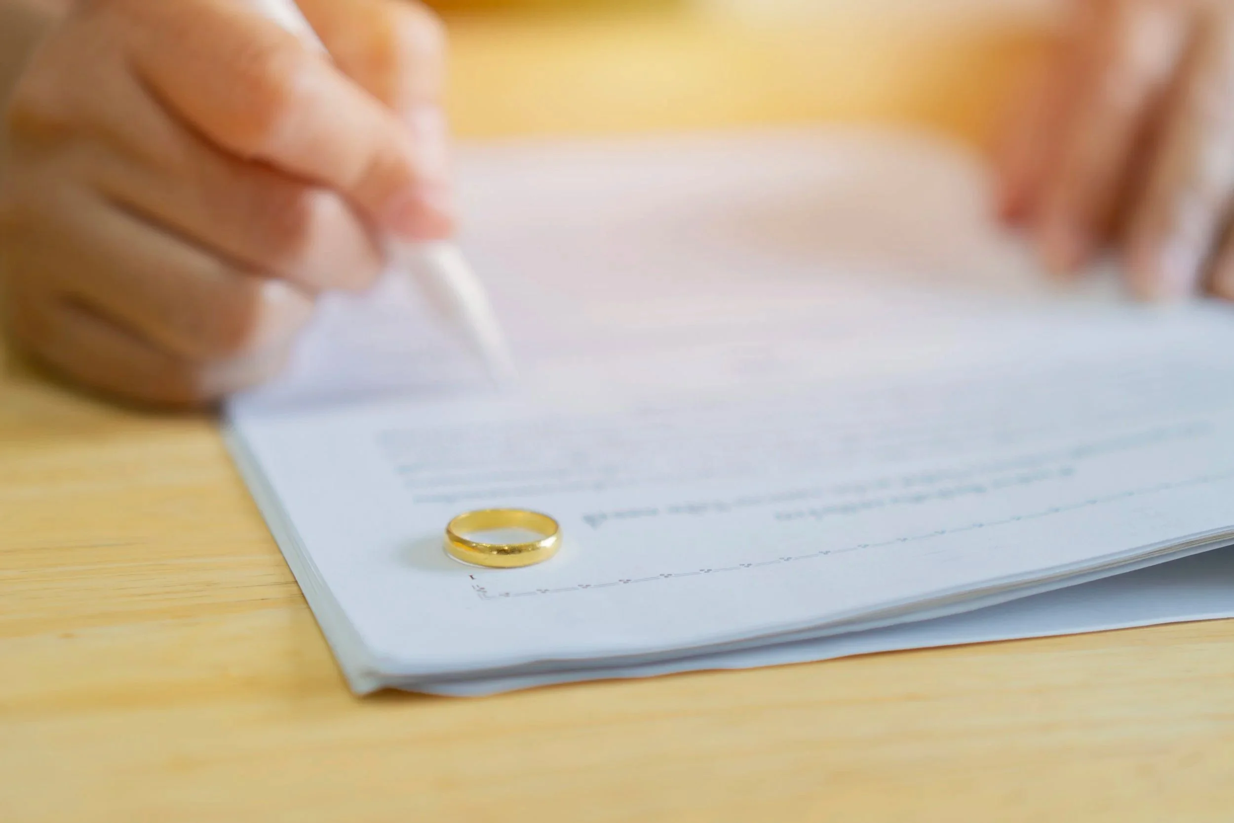 A person holding a pen above a lined paper with a gold wedding ring placed on the paper, on a wooden surface.