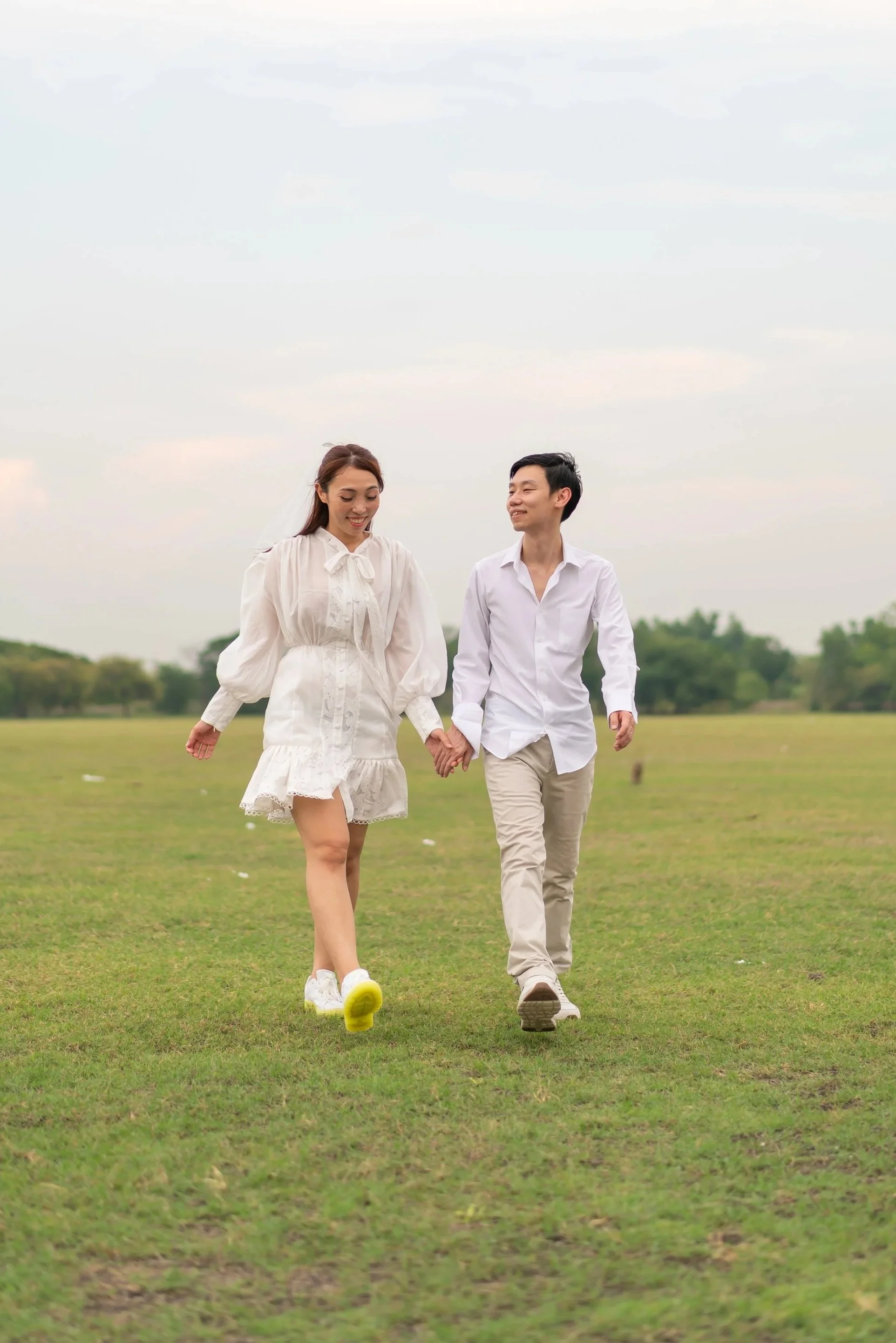 A couple walking hand-in-hand across a grassy field, smiling and enjoying each other's company on a cloudy day.