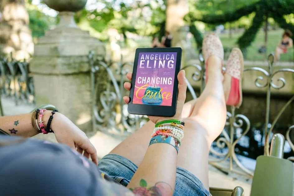 Person sitting outdoors on a bench or ledge, holding a tablet displaying a book cover titled 'Changing Course' by Angelica Eling. The person is wearing colorful bracelets and has tattoos on their arms. Their legs are stretched out, and they are wearing light-colored shoes. The background features decorative ironwork, lush greenery, and people sitting in the park.