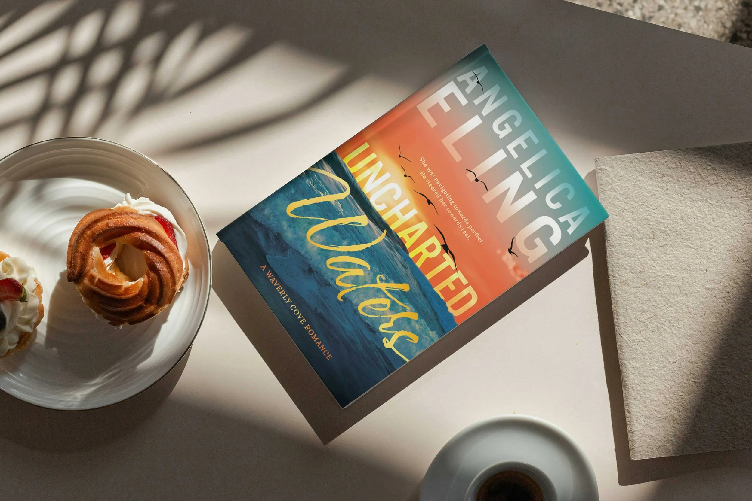 A mug of coffee, a white bowl with pastry and whipped cream, a book titled 'Uncharted Waters' and a plate with a pastry are arranged on a light-colored surface with shadows cast by nearby leaves.