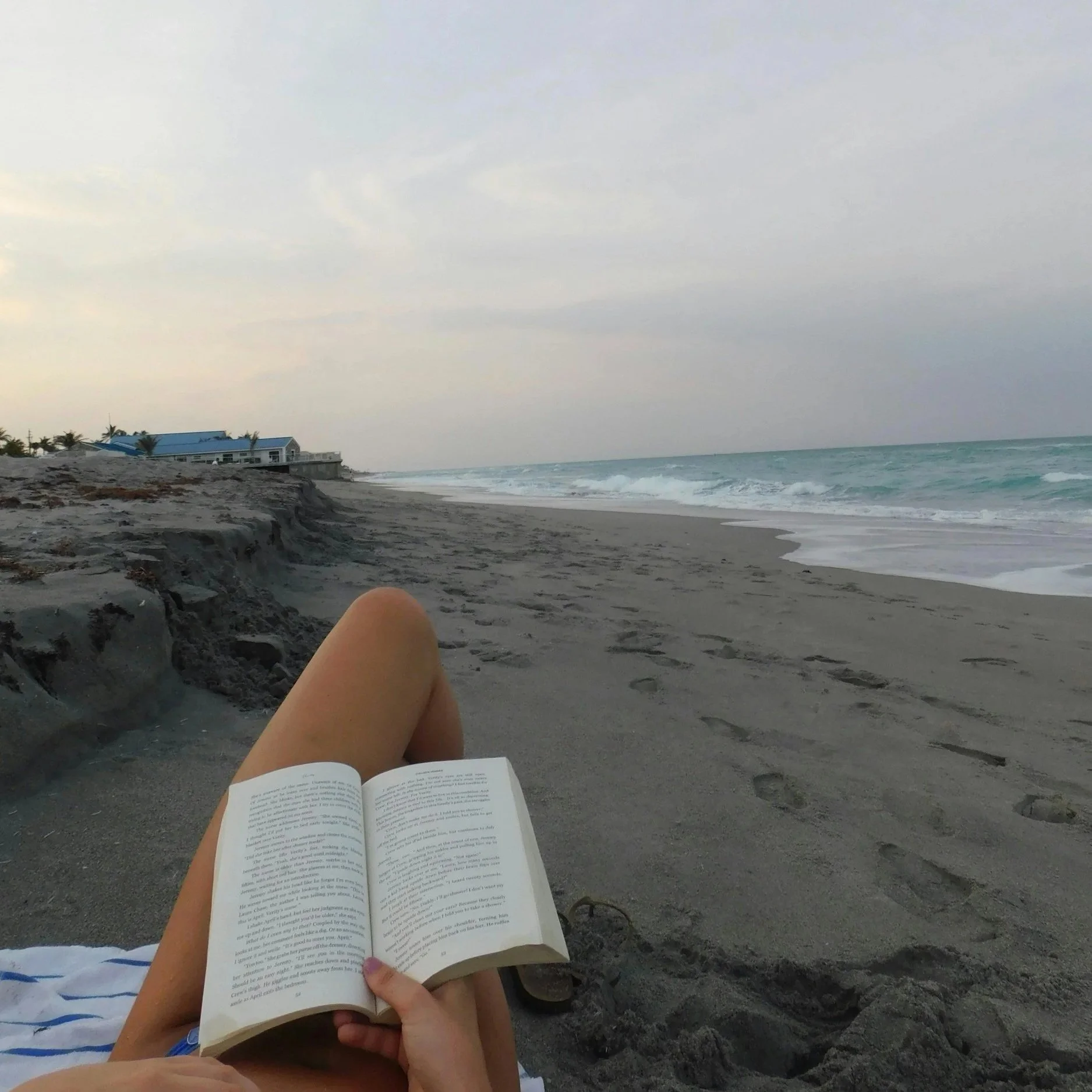 A person sitting on a sandy beach reading a book, with the ocean waves and a cloudy sky in the background. They are wearing a towel with blue and white stripes and have bare legs.