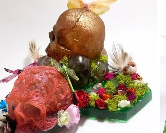 Mixed media sculpture titled 'Beauty in Death' by Breanna Wilbur, featuring gold-painted skulls, vibrant flowers, feathers, a butterfly, and decorative elements arranged on a platform.