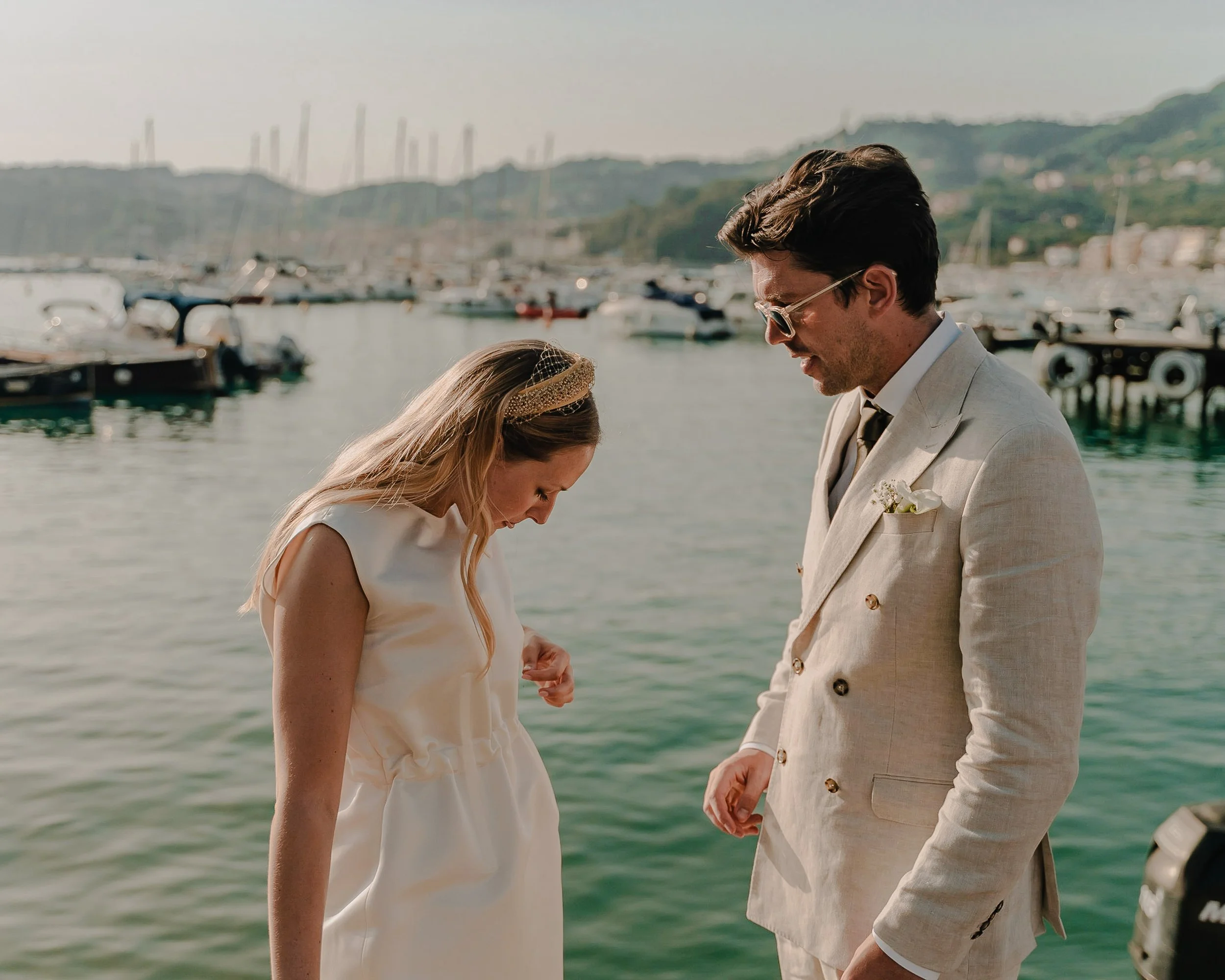 Couple in elegant attire by marina with boats and hills in background.