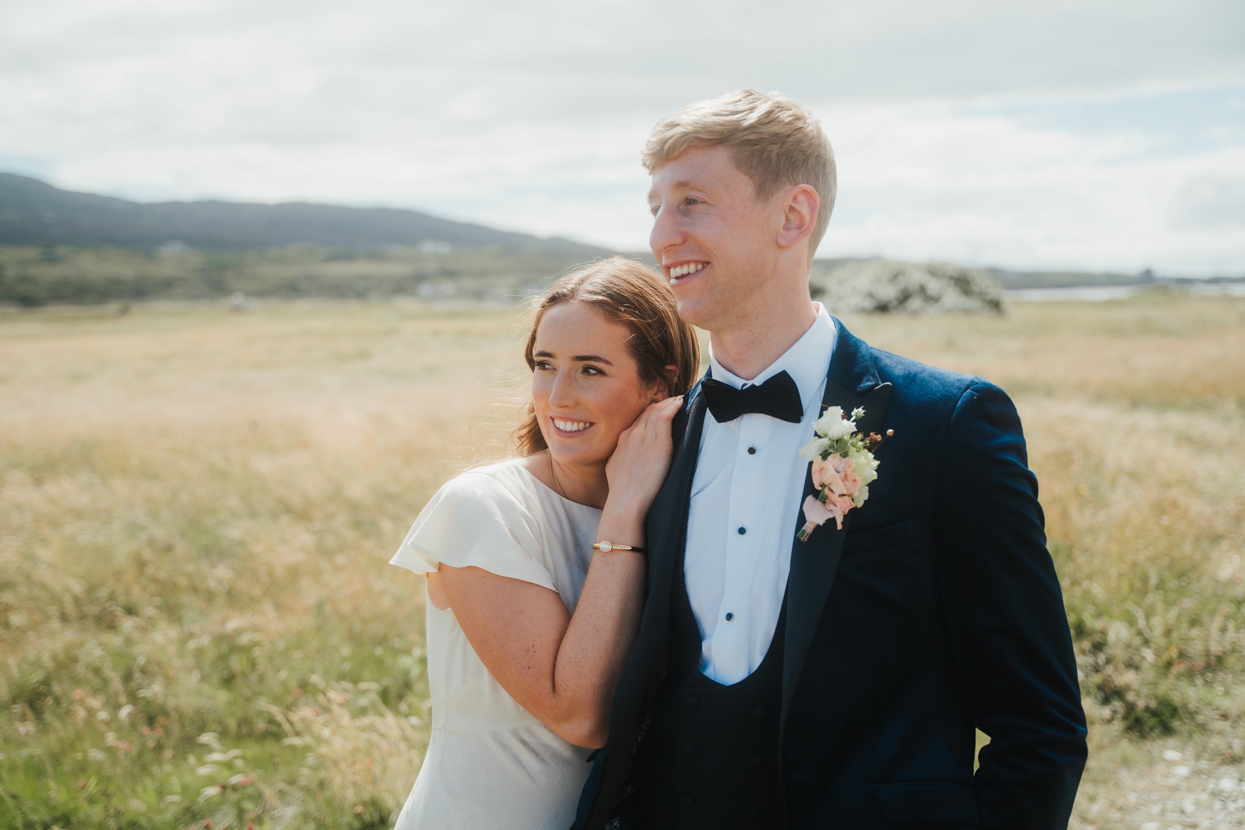 Ein lächelndes, formell gekleidetes Paar steht auf einem offenen Feld. Die Frau in einem weißen Kleid lehnt sich an den Mann in einem dunklen Anzug mit einer Boutonnière. Im Hintergrund sind Hügel und ein bewölkter Himmel zu sehen.