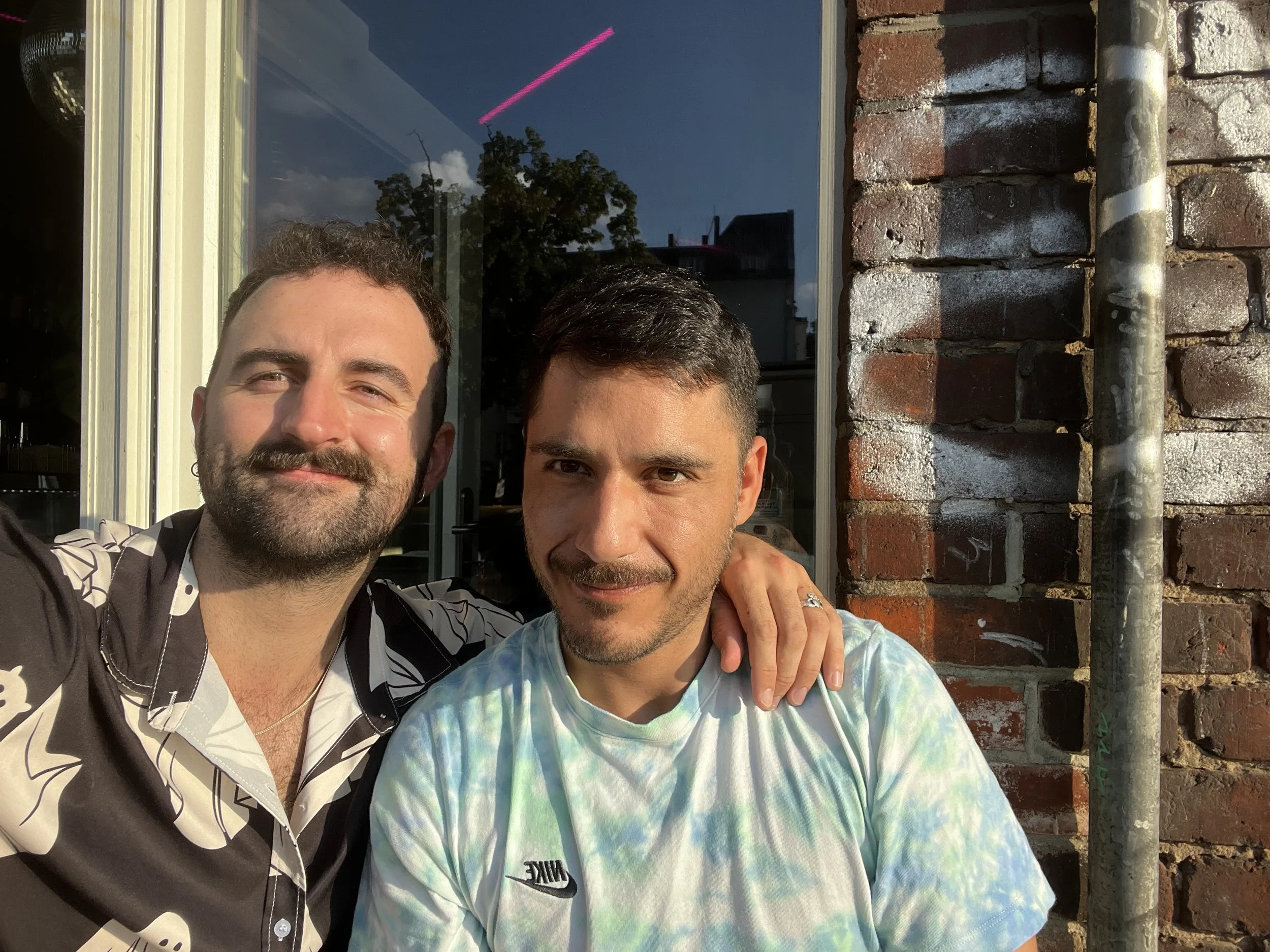 Two people smiling, one wearing a black shirt with white ghost patterns, the other in a light blue tie-dye shirt, standing outdoors with a brick wall and a window in the background.