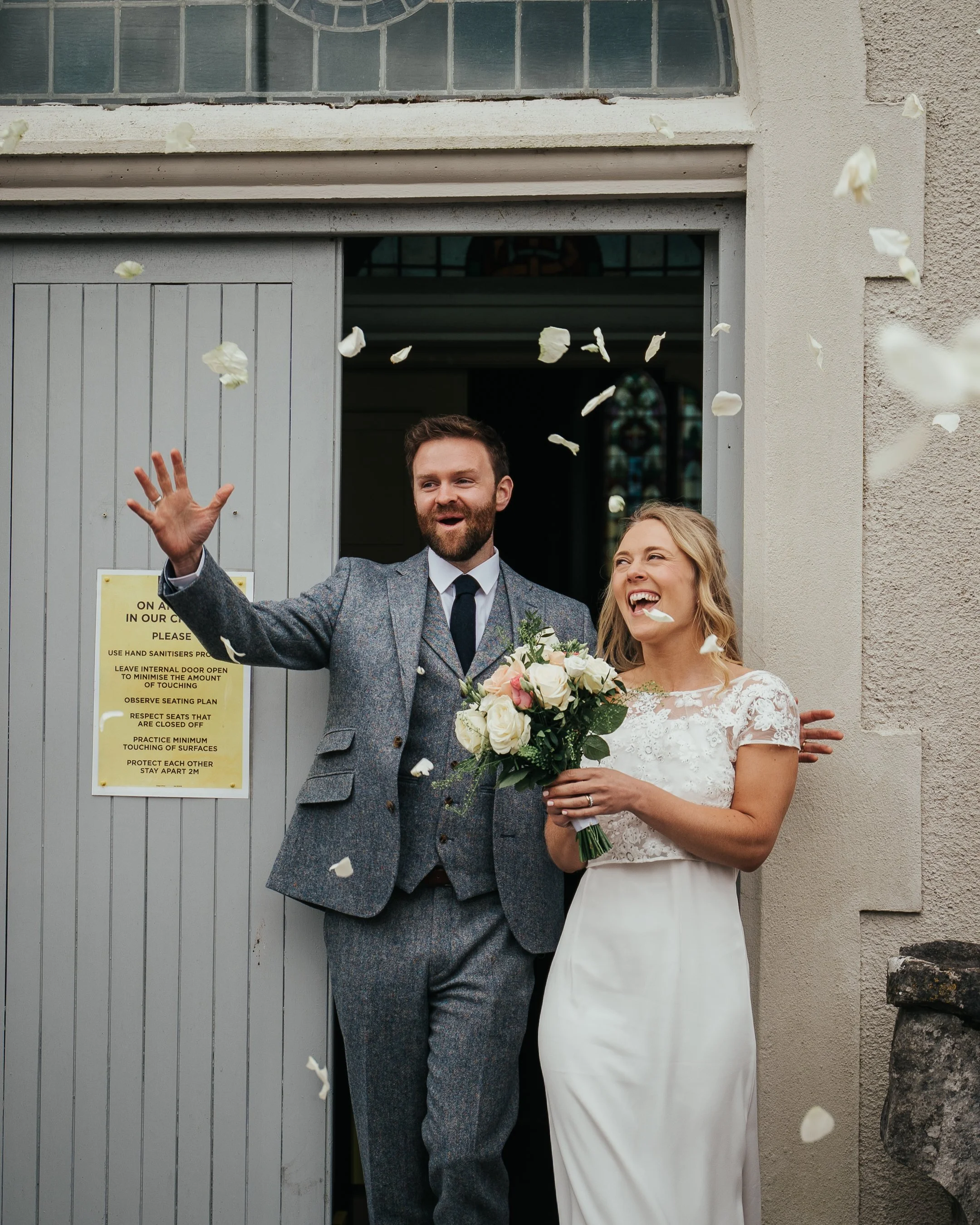 Bride and groom leaving a building, smiling and celebrating, with the groom throwing confetti petals. The bride holds a bouquet of flowers, wearing a white wedding dress, and the groom is in a gray suit.