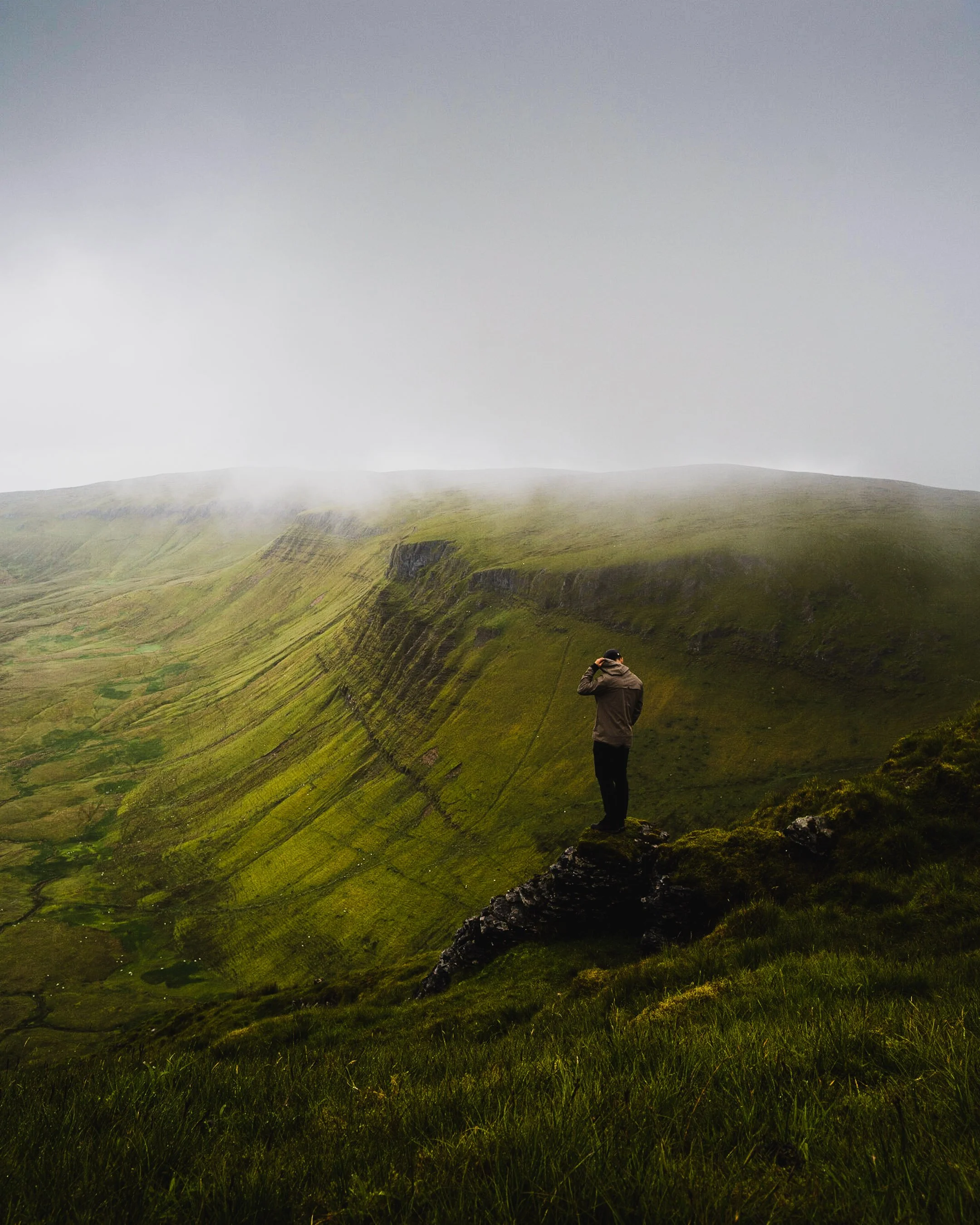 Person standing on a grassy cliff edge overlooking a foggy, mountainous landscape with a sweeping green valley.