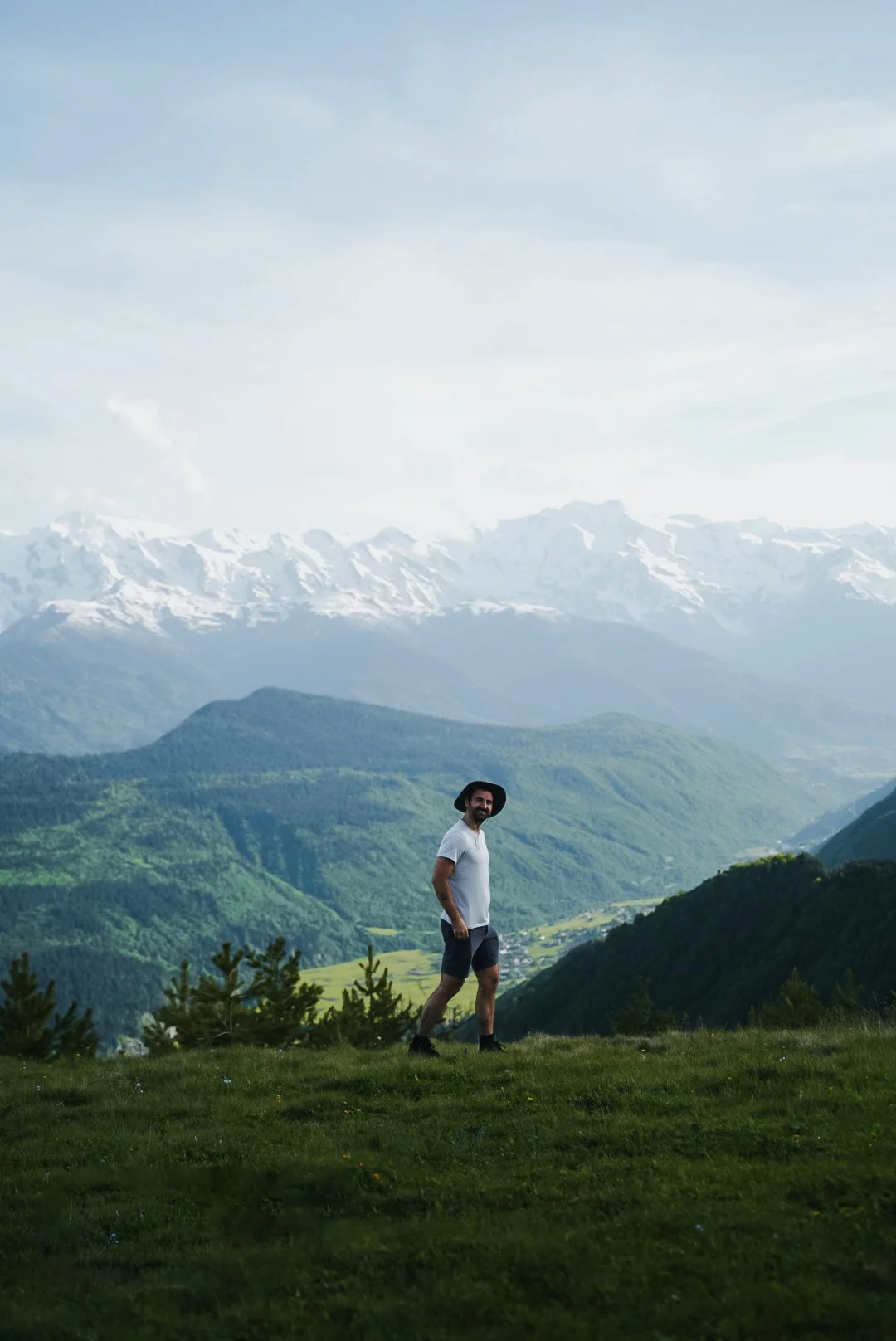Man in hat and shorts walking on a grassy hill with mountain range in the background.