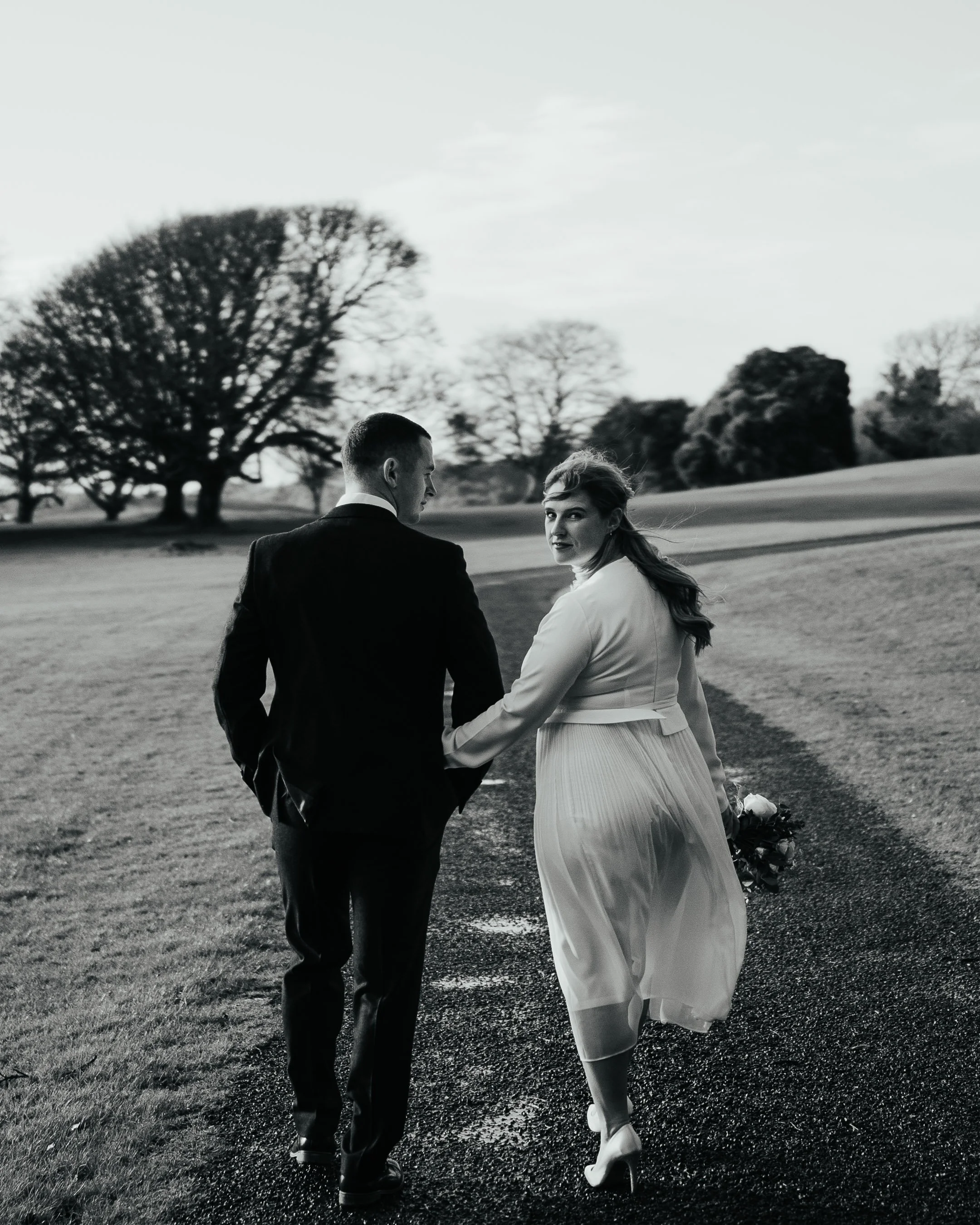 A black and white photo of a couple walking hand in hand on a path, with the woman holding a bouquet. They are outdoors, surrounded by grass and large trees in the background.