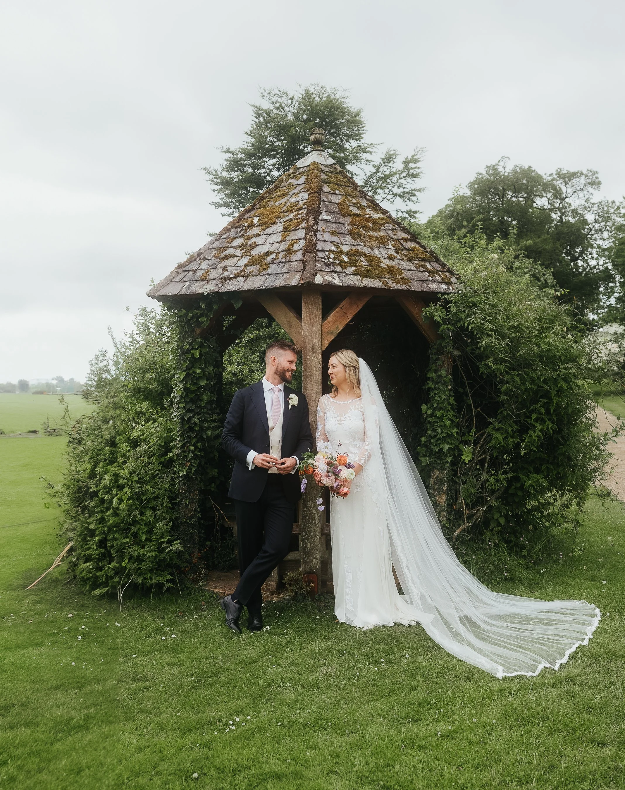 Bride and groom standing under a wooden gazebo in a garden setting.