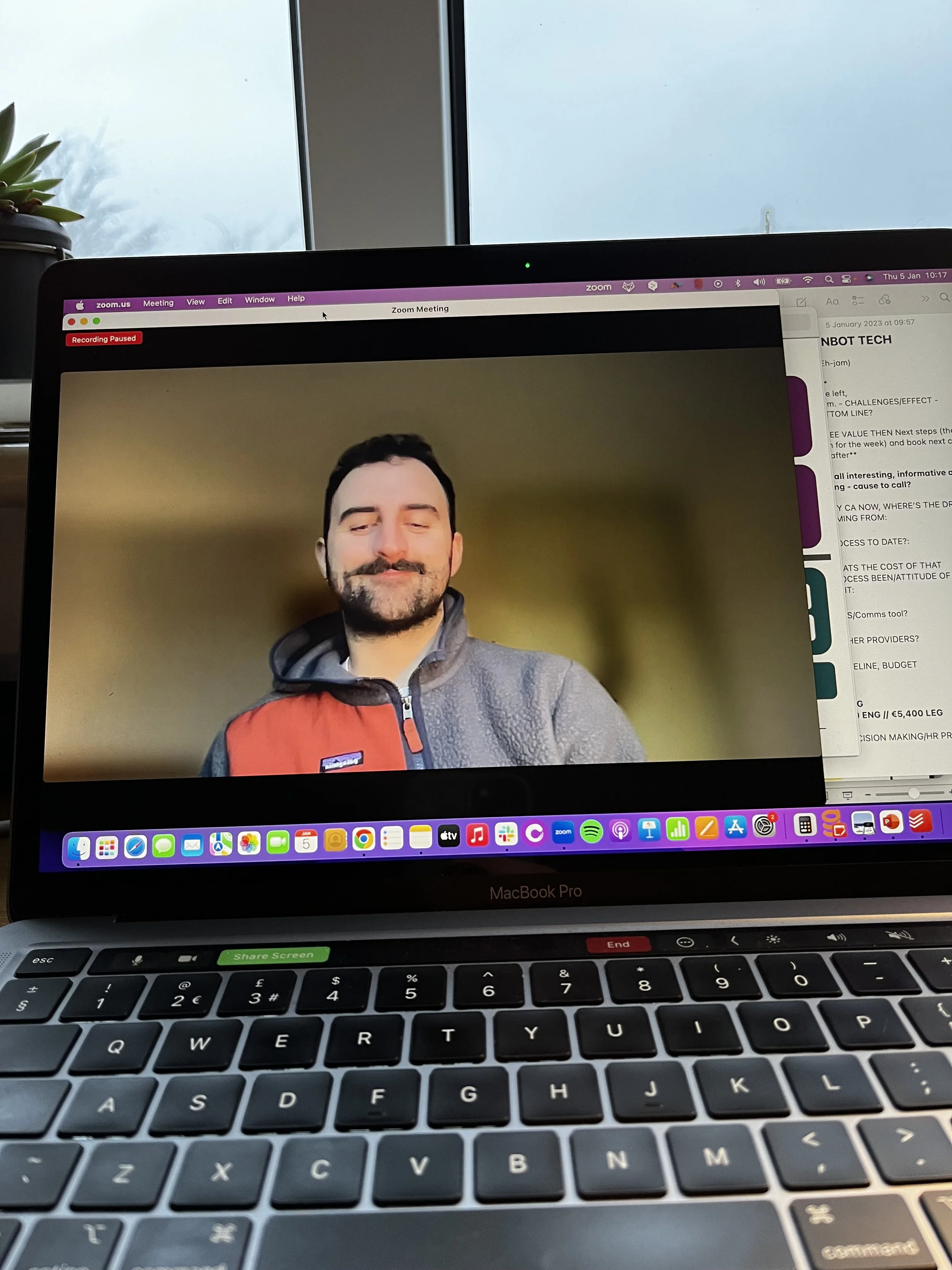 Laptop screen displaying a paused Zoom meeting with a person in a gray and orange jacket, in front of a window with overcast sky.