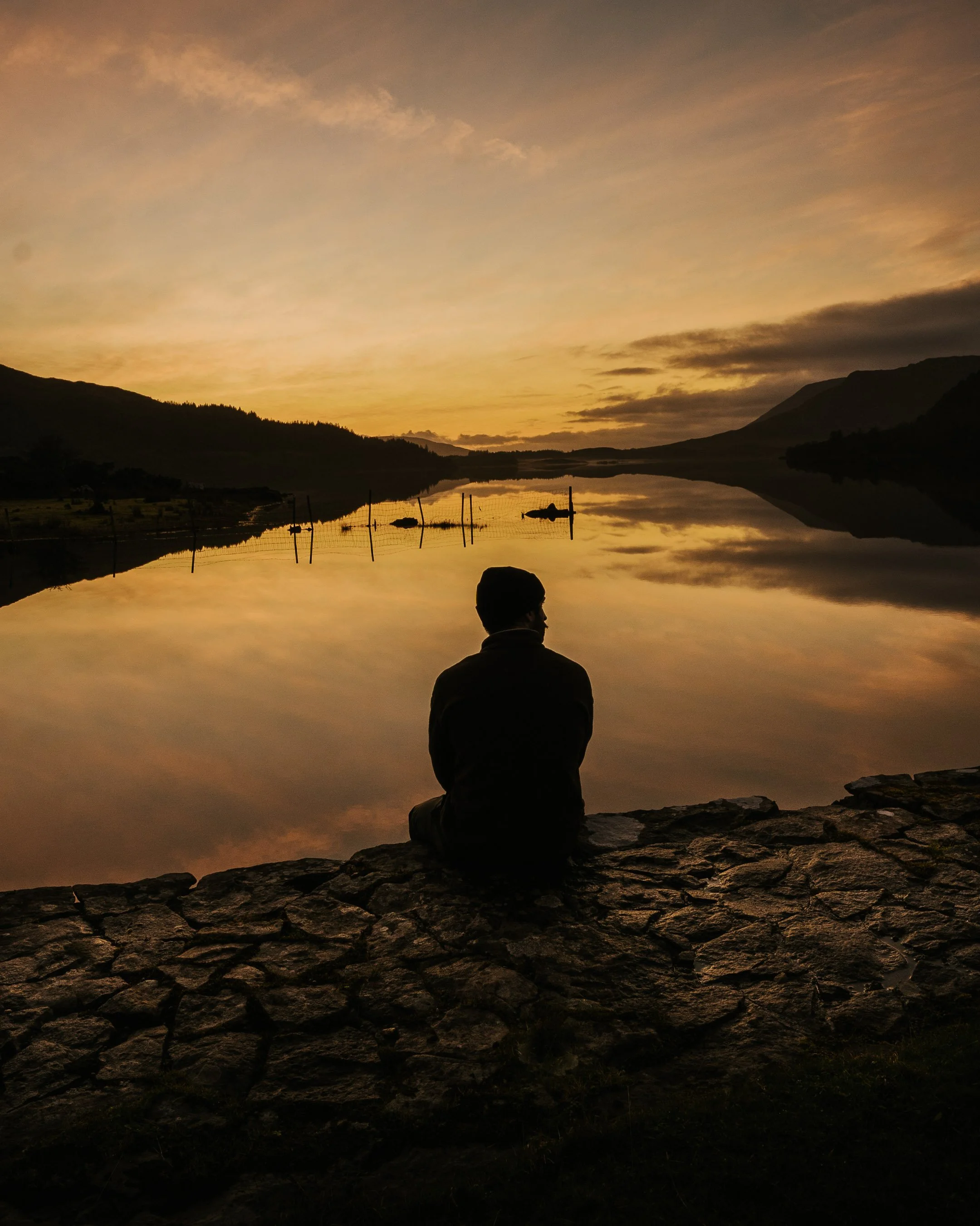 Silhouette of person sitting on a rocky surface by a calm lake during sunset, with mountains and trees in the background.