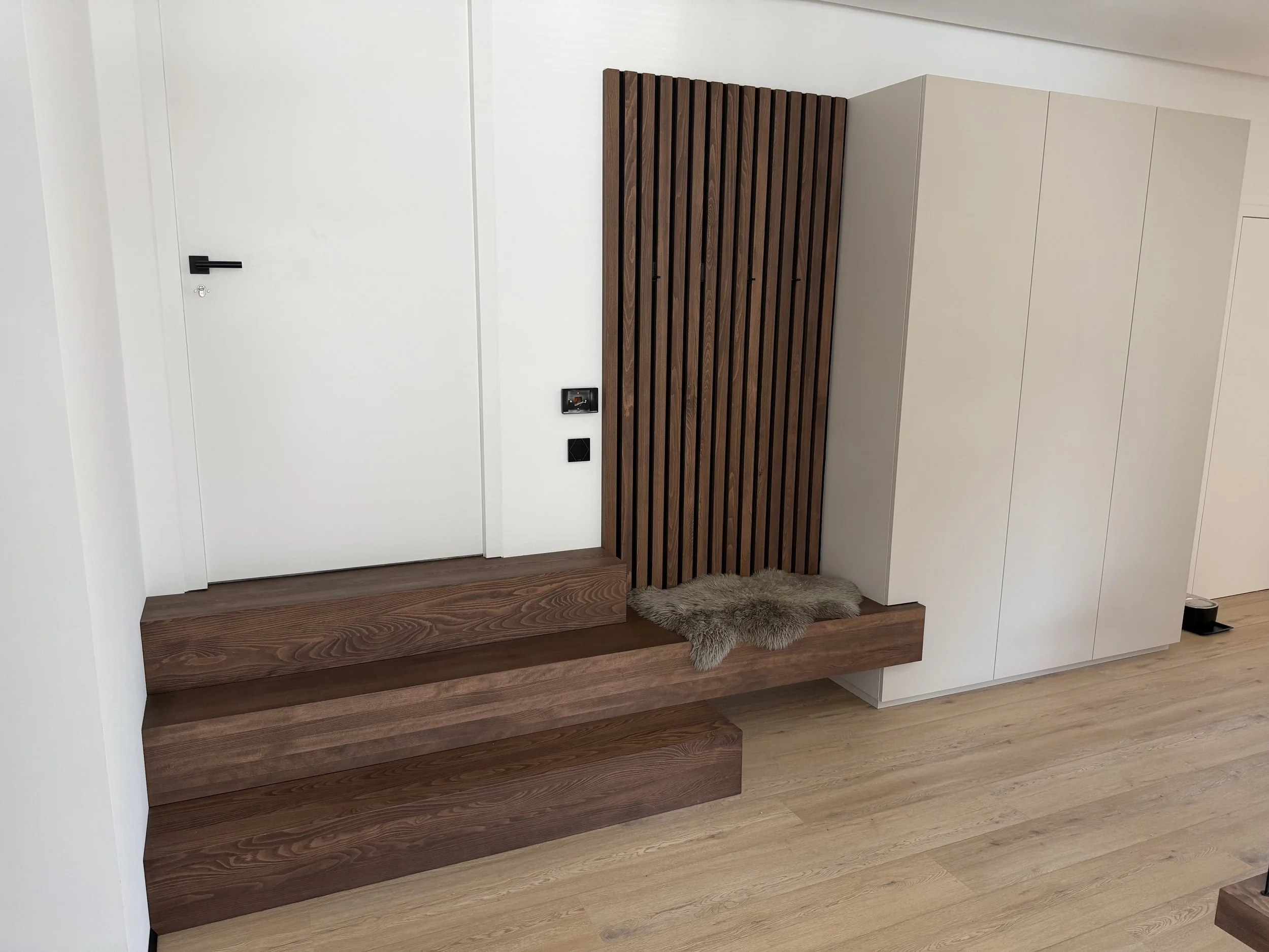 Innenaufnahme eines Hauses mit weißen Wänden, einer Treppe aus Holz, einem Holz-Armlehnen-Element an der Wand und einem kleinen Teppichmuster auf dem Boden.