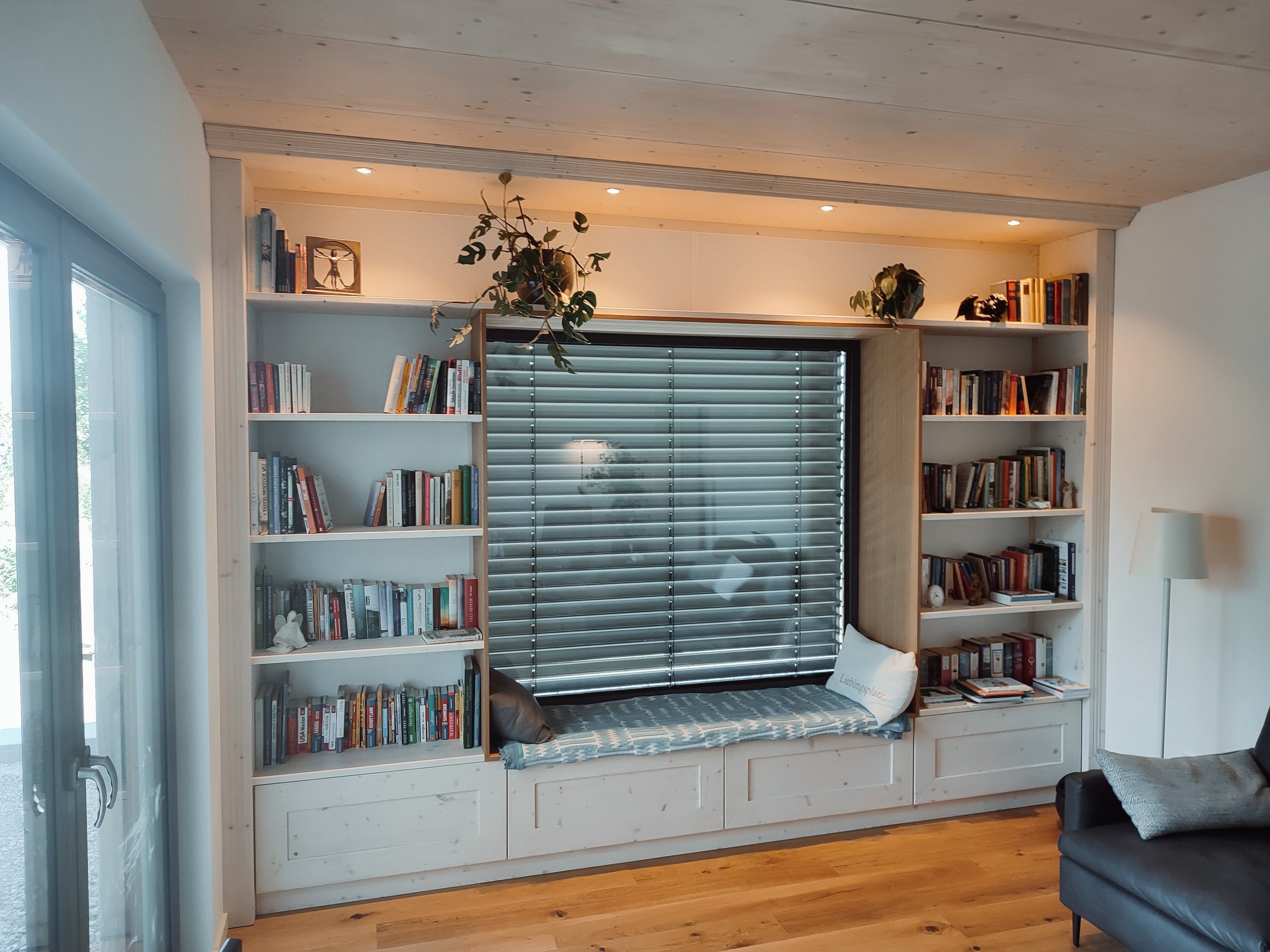 Ein Wohnzimmer mit weißen Bücherregalen, einem großen Fenster mit Rollläden, einer Bank mit Kissen und einem Ohrensessel.