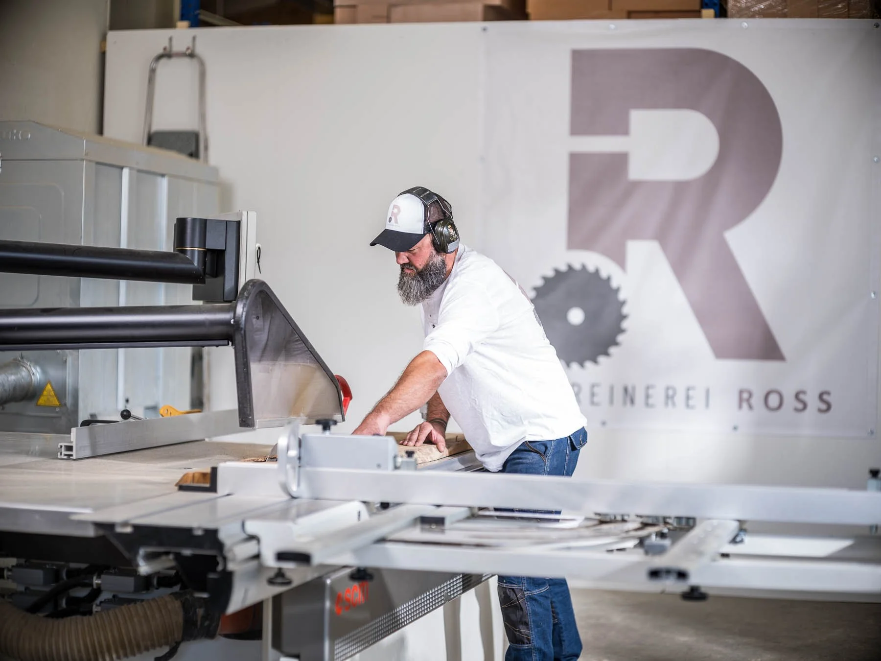 Mann arbeitet an Holzbearbeitungsmaschine in einer Werkstatt, im Hintergrund ein Banner mit einem großen Logo einer Säge und dem Text 'Reinerei Ross'.