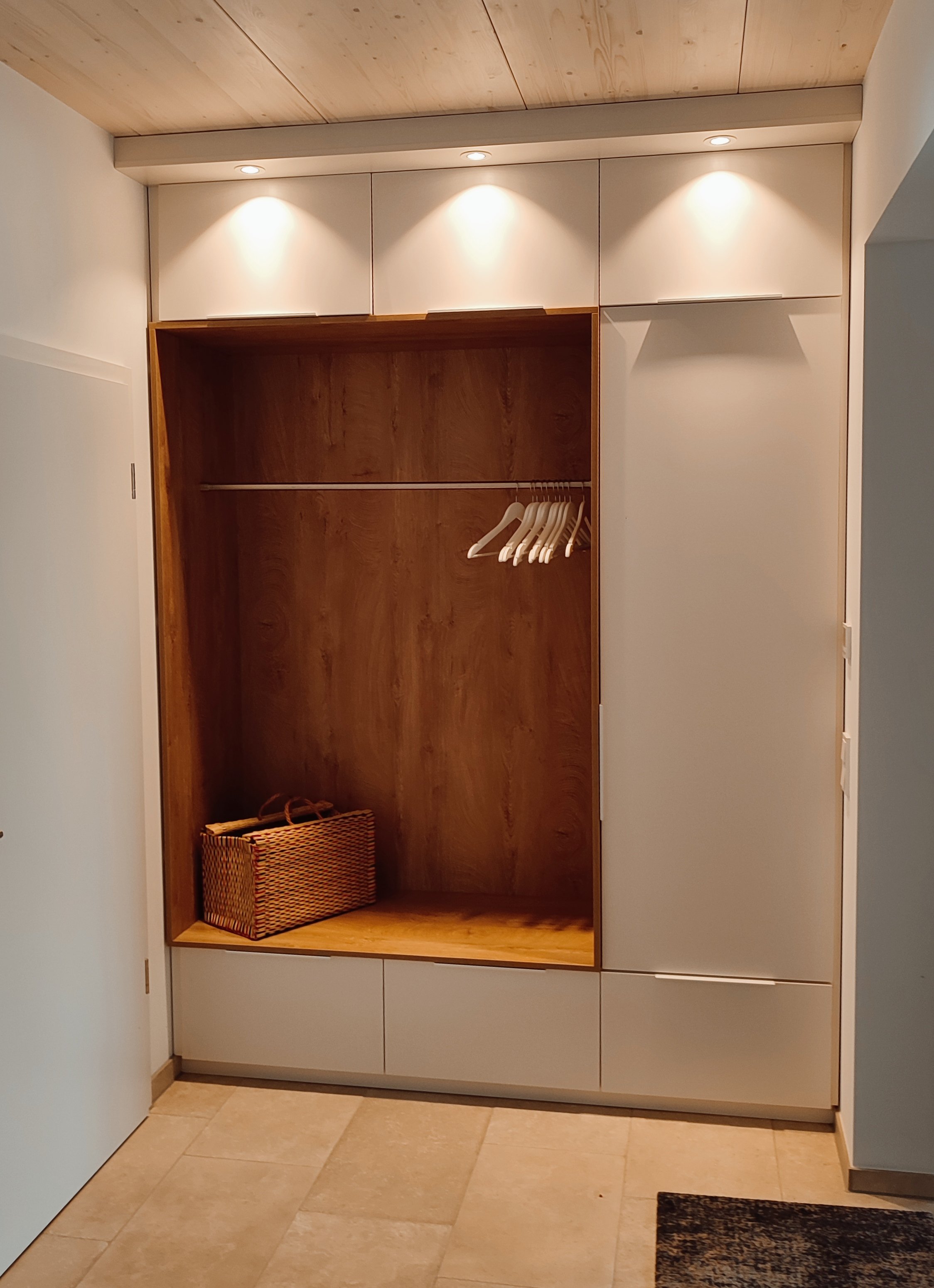 Offene Kleiderschrank mit einem kleinen Korb und mehreren weißen Kleiderbügeln, beleuchtet durch drei eingebaute Deckenleuchten.