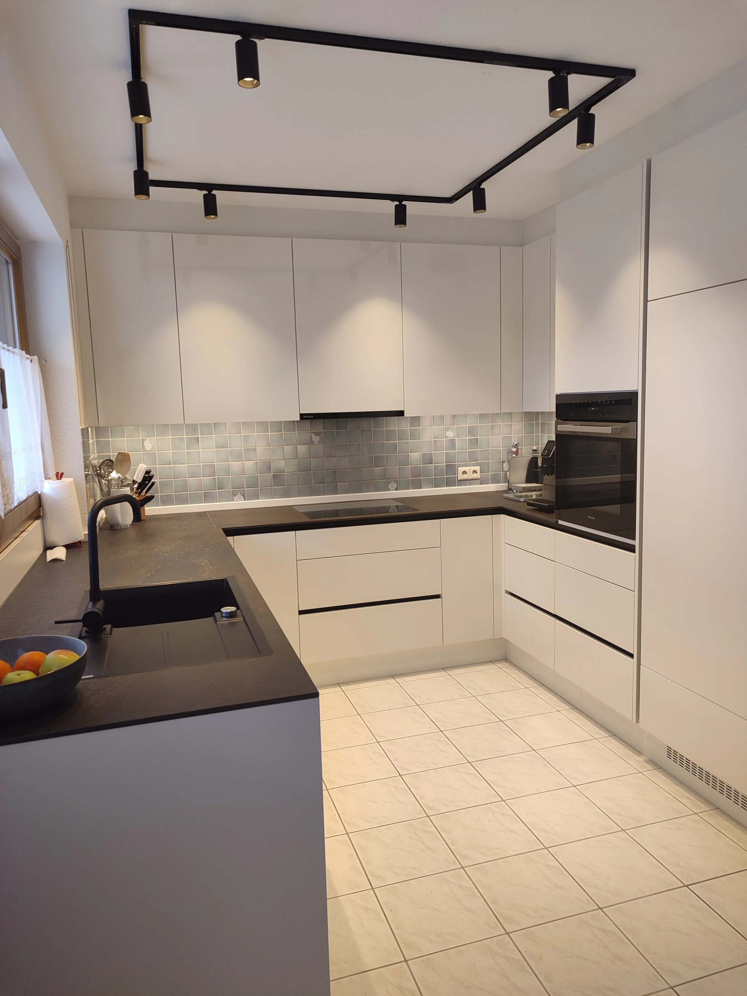 Moderne weiße Küche mit schwarzem Arbeitsbereich, Backofen, Spüle, Geschirr und Fruchtenschüssel, amplem Stauraum und dunkler Deckenbeleuchtung.