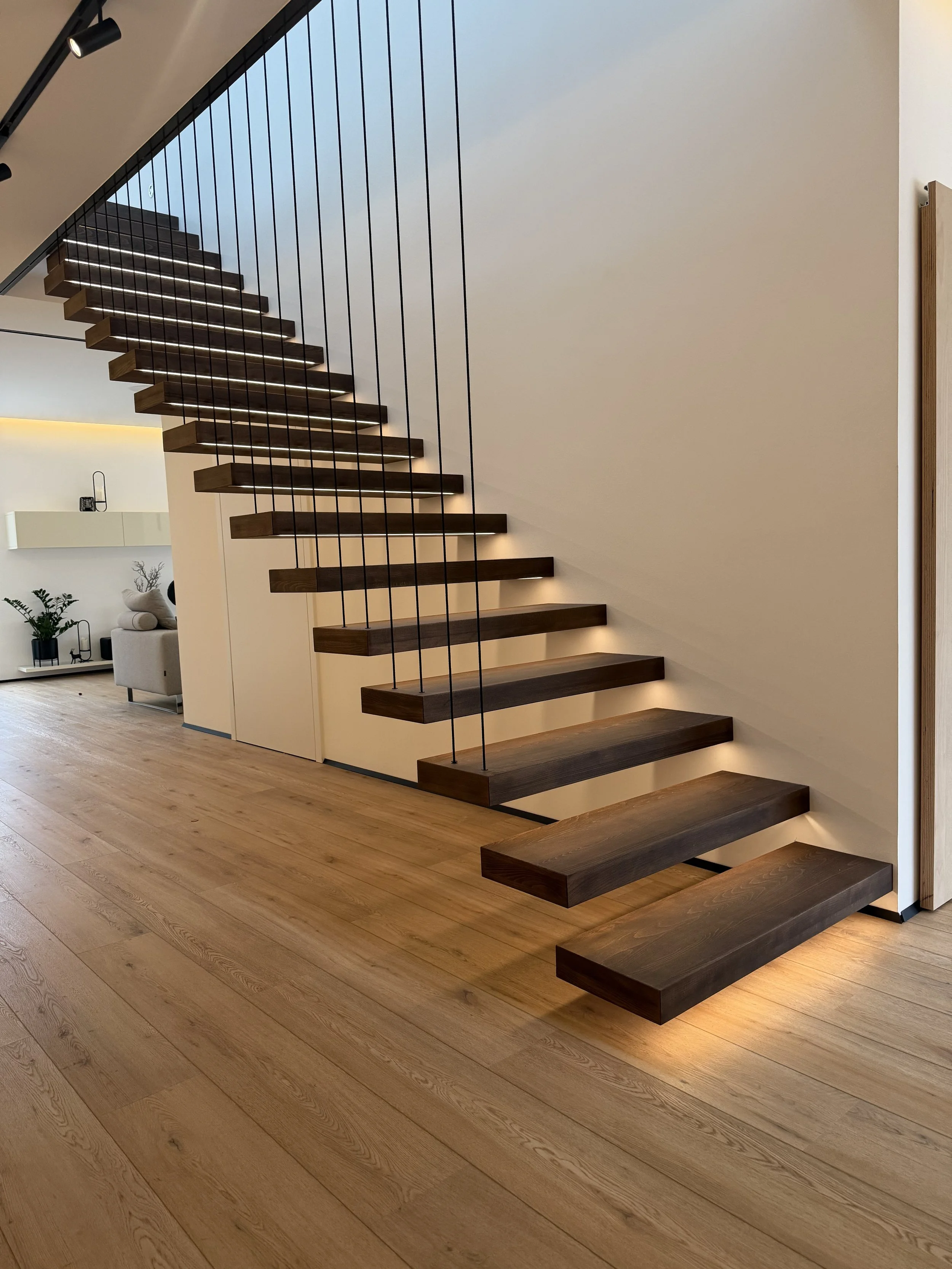moderner, offener Treppenaufgang mit dunklen Holzbrettern und schwarzen Metallseilen, helle Wände und Holzfußboden