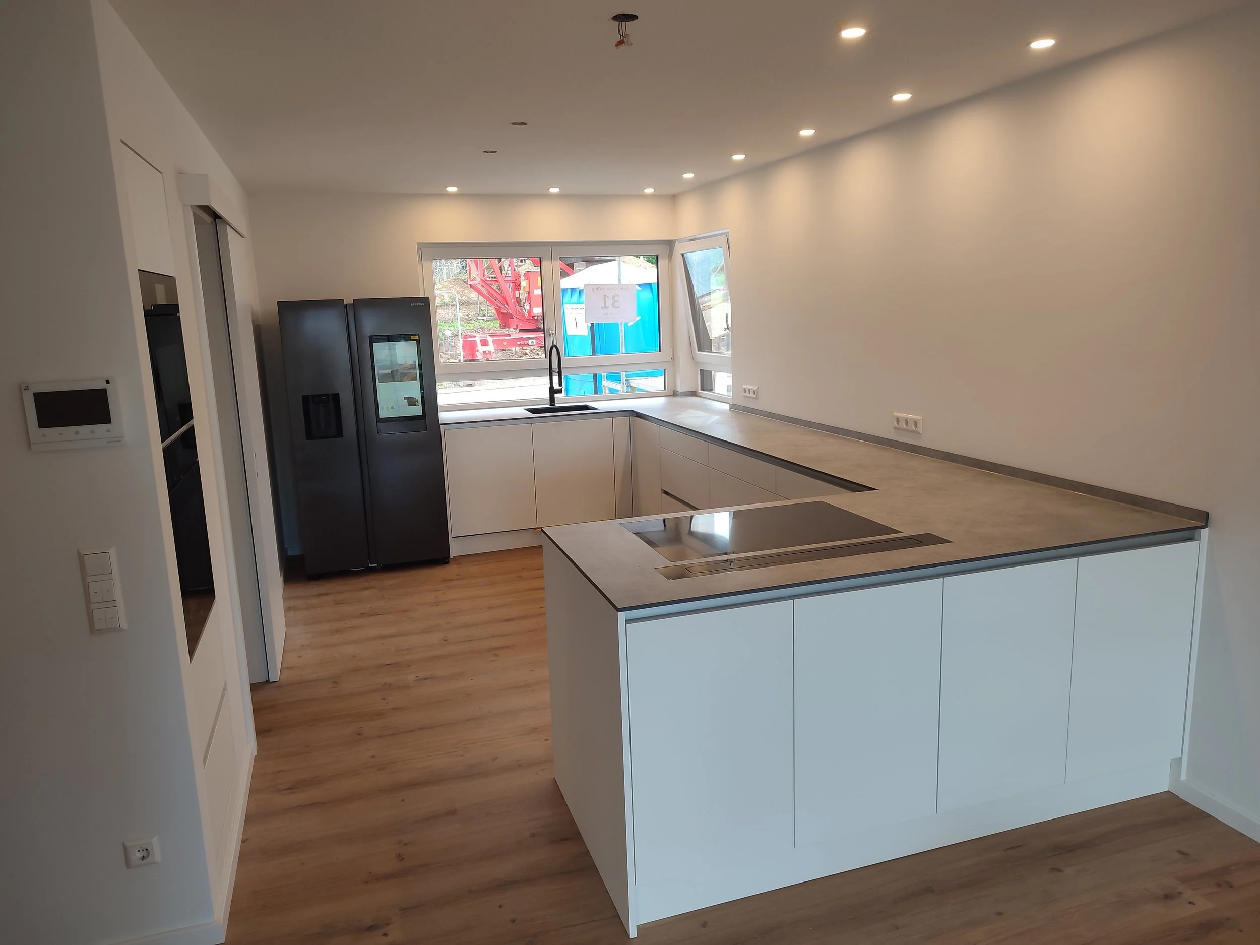 Moderne, helle Küche mit weißen Schränken und dunklem Stein- oder Granit-Arbeitsplatte. Es gibt große Fenster, die viel Tageslicht hereinlassen, und einen Kühlschrank. Die Küche befindet sich im Bau, da die Deckenleuchten noch installiert werden.