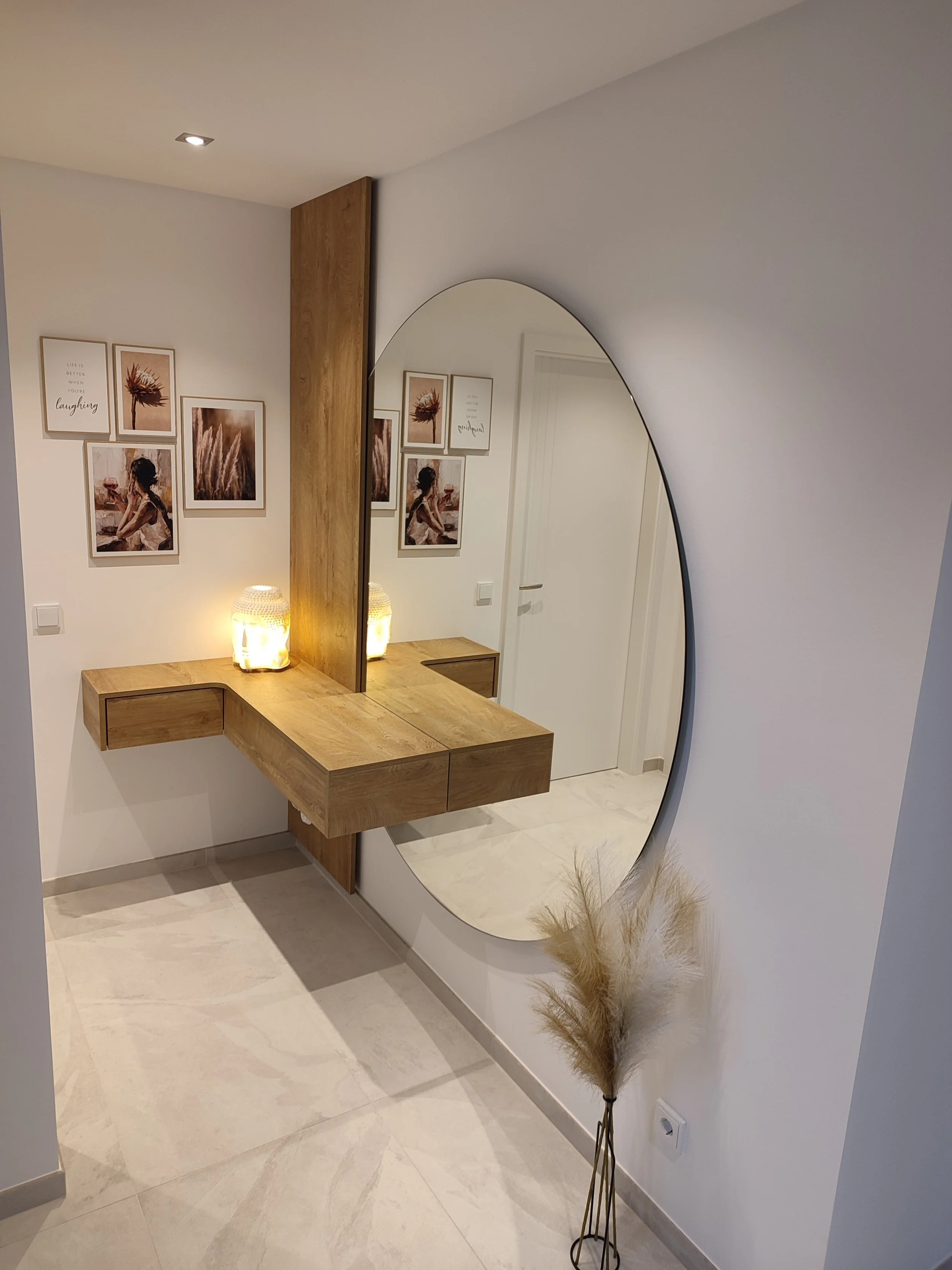 Ein modernes Flur mit einem großen ovalen Spiegel an der Wand, einem Wandregal aus Holz, dekorativen Lampen, Wandbilder mit Naturmotiven und einem Deko-Strauß mit Trockenblumen.