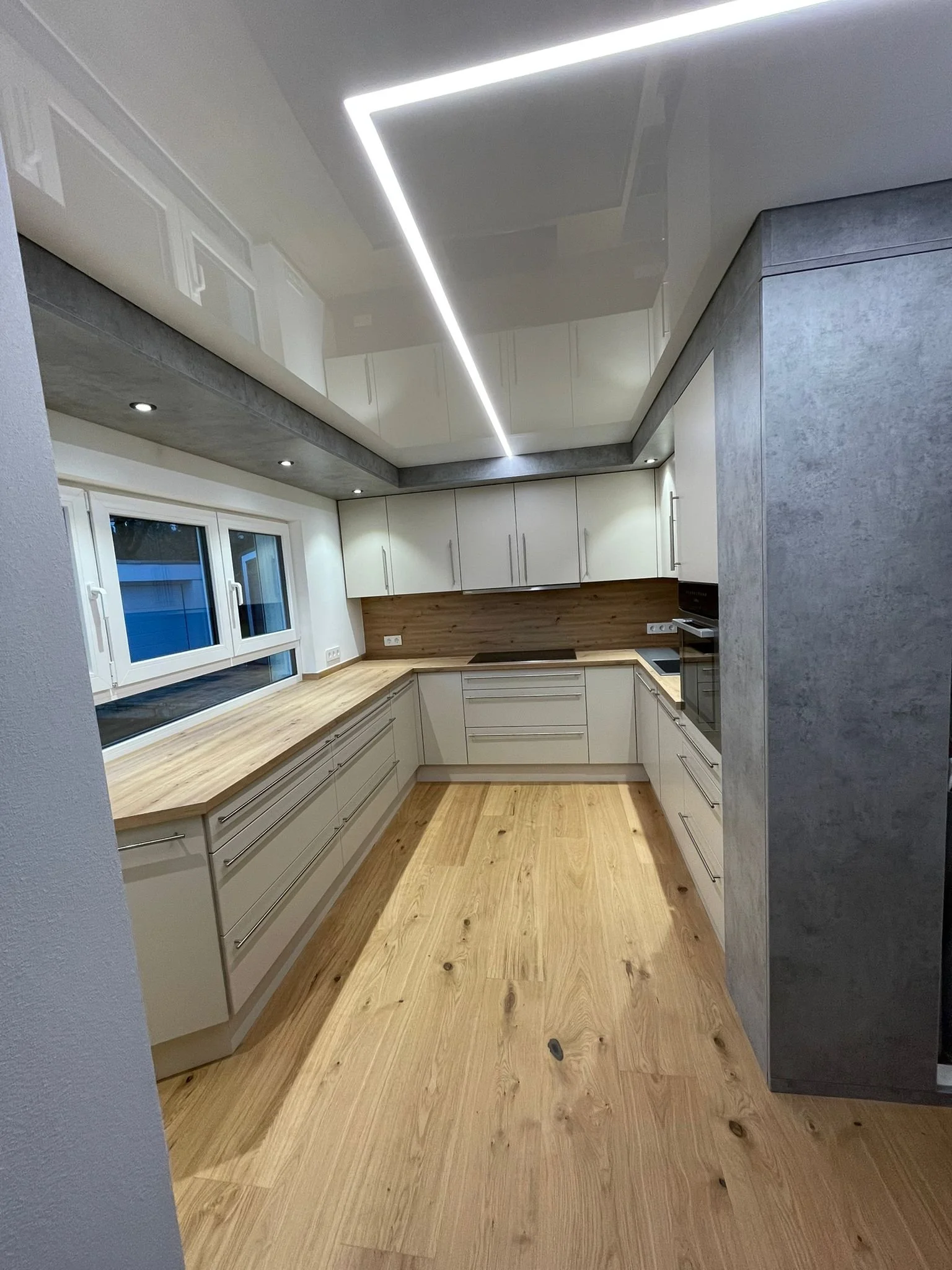 Moderne Küche mit weißen Schränken, Holz-Arbeitsplatten, großen Fenstern und Holzfußboden, beleuchtet durch eine moderne LED-Deckenleuchte.